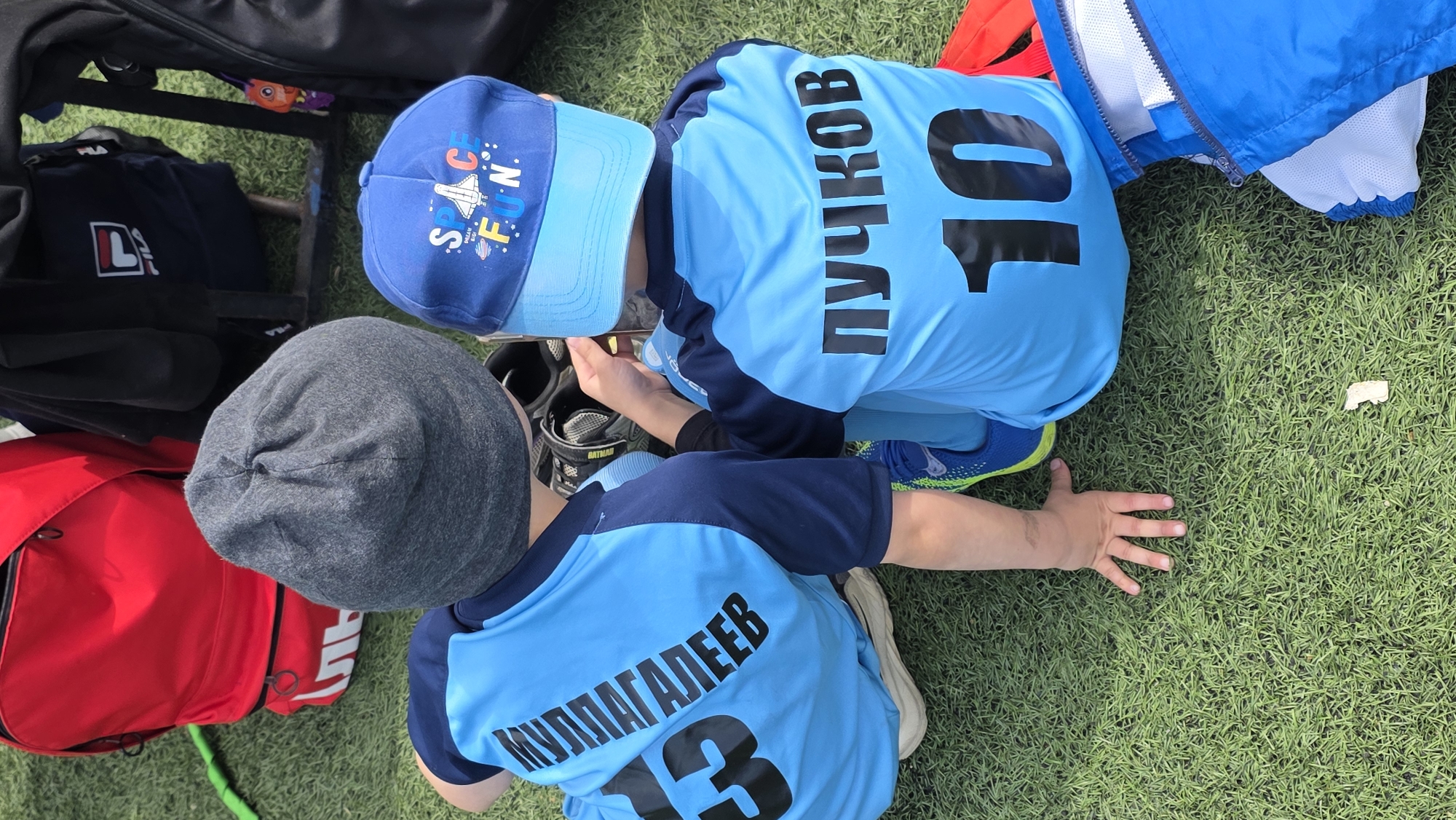 A photograph shows two children wearing matching light blue jerseys with dark blue sleeves, dark lettering, and numbers on the backs, resting on what appears to be a green artificial turf surface outdoors. The child on the right wears a blue cap with the words "SPACE FUN" on it and the number "10" displayed prominently on the back of the jersey. To the left, another child is wearing a gray beanie and a jersey with the number "13" on the back. The name on the back of the child with the number 13 jersey says "МУЛЛАГАЛЕЕВ". The name on the back of the child with the number 10 jersey says "ПУЧКОВ". The children appear to be interacting with an electronic device, likely a phone or a small tablet, which they are holding between them. Several bags and a jacket are visible around them, suggesting a sports setting.