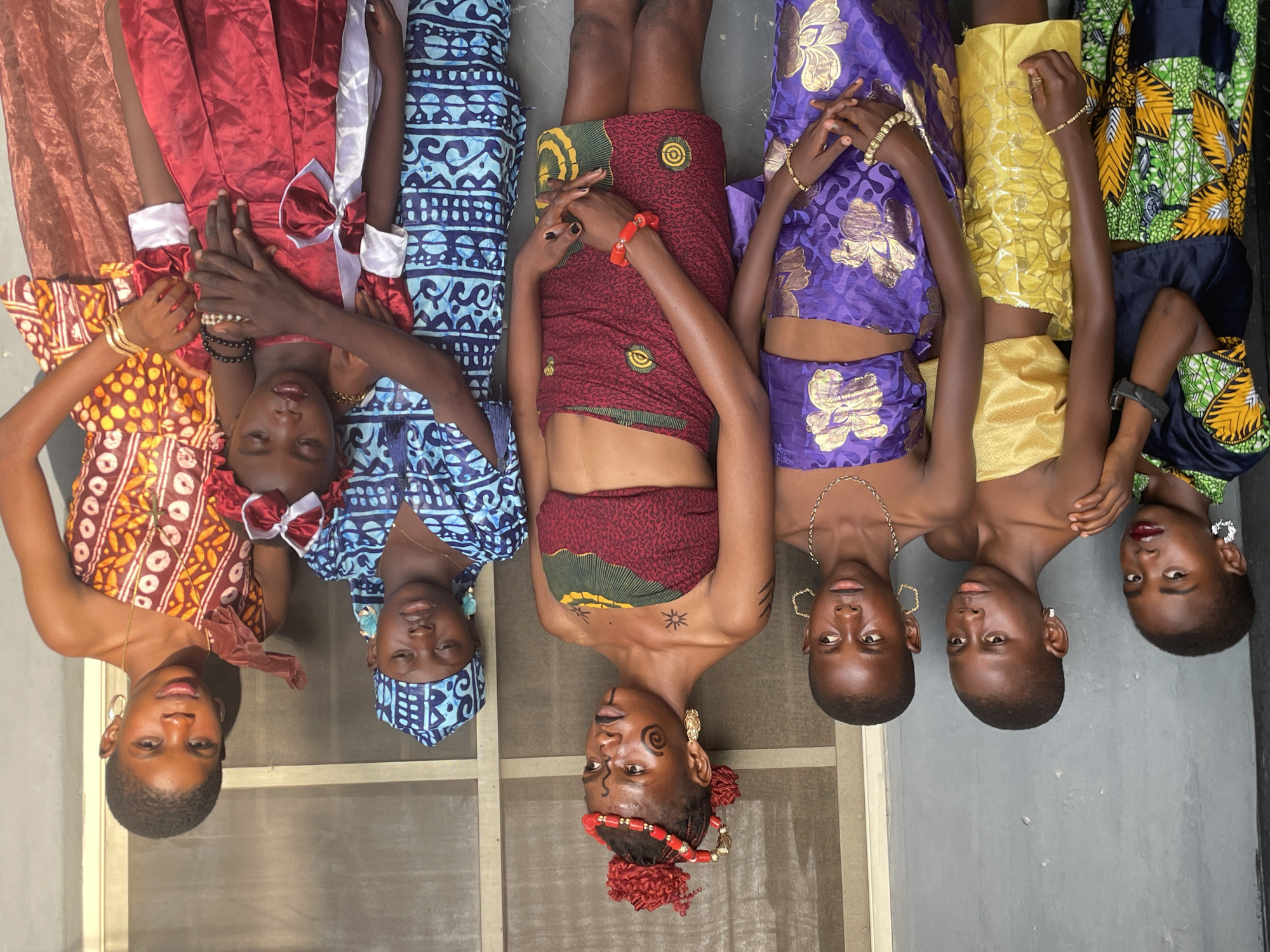 Six young women are depicted lying side-by-side on a light-colored surface, posing for a photograph in what appears to be an indoor or sheltered outdoor setting in Badagry, Nigeria. They are arranged horizontally across the frame, with their heads aligned and facing upwards towards the viewer.
Each woman is dressed in distinct, colorful traditional or festive attire, showcasing a variety of patterns and fabrics. From left to right, the garments include an orange and brown patterned dress, a red dress with white bows and trim, a maroon patterned skirt and top, a purple garment with gold floral accents, a yellow top and skirt, and a green and black patterned outfit.
The women are adorned with various accessories: bracelets (gold, beaded, and a watch), necklaces (gold chains), and earrings (hoops, decorative gold pieces). The central figure is particularly notable, wearing an elaborate red beaded headpiece, large gold earrings, and intricate black body paint or markings on her face, neck, and chest, suggesting a cultural celebration or ceremonial context.
The lighting is natural and bright, indicating it is daytime. Their facial expressions are generally serene and composed, consistent with posing for a portrait. The background is simple, featuring a plain wall or doorframe. No visible text is present in the image. The scene captures a moment of cultural pride and communal display.