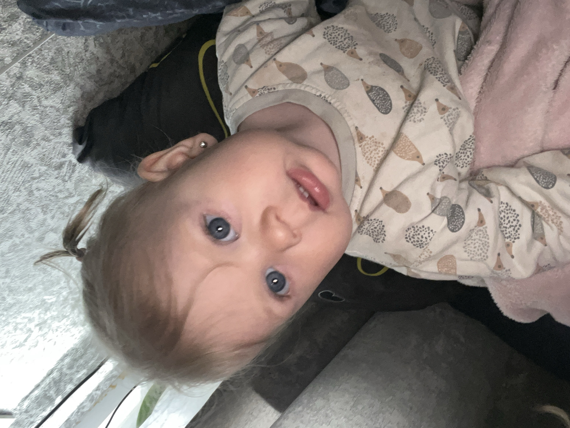A young baby or toddler with light blonde hair and striking blue eyes is depicted in a close-up, sideways-oriented photograph. The child is lying down, looking directly at the camera with wide eyes and slightly parted lips, conveying a neutral to curious expression. A small, silver stud earring is visible in the child's left earlobe. Their light-colored hair is styled with a small ponytail on the left side of the head.
The baby is dressed in a cozy long-sleeved onesie or pajama top adorned with a repeating pattern of cartoon hedgehogs in various shades of brown and grey. They appear to be resting on a dark fabric surface, possibly a pillow or part of a bed, with a soft, plush pink blanket or fabric visible to their right. The background on the left features a light grey, textured surface, resembling a rug or carpet. The indoor lighting is soft and ambient. There is no visible text in the image. The image was captured in Фоки, Russia.
