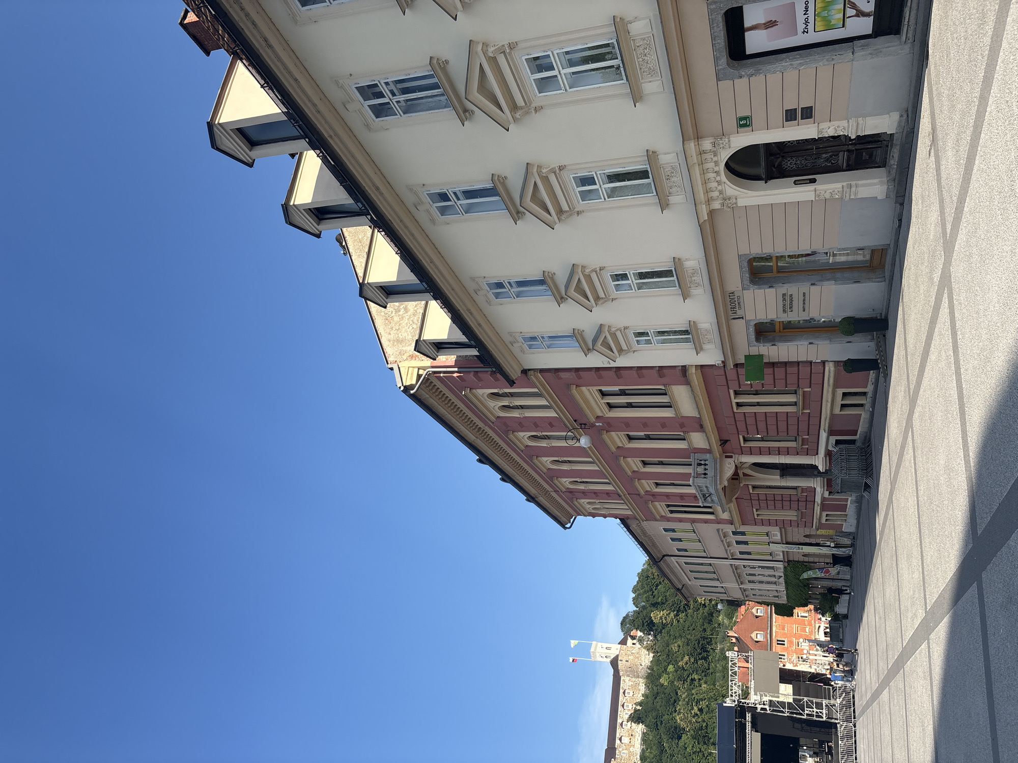 A wide-angle, low-angle shot captures the exterior of buildings and a bright blue sky. The buildings are tall and appear to be historic, with multiple stories featuring windows and ornate architectural details. The facades are a mix of light beige and pinkish-red. Some windows are rectangular with white frames, while others have elaborate triangular decorative elements above them. Dormer windows are visible on the upper sections of the buildings, protruding from the roofline and angled towards the sky.
In the distance, on a hill, a castle structure with flags is visible, surrounded by lush green trees. In the foreground, a paved plaza is visible with a stage set up, indicating an outdoor event or performance. The lighting suggests it is daytime, likely mid-morning or afternoon, with clear, sunny weather. The scene appears to be a bustling urban environment, possibly a city square or promenade, with a mix of historical architecture and modern event staging.
Text is visible on a sign on one of the buildings: "AFRODITA", "LJUBLJANA CENTER", and "SPLETNA TRGOVINA". Another sign displays "Živjo, Neo".