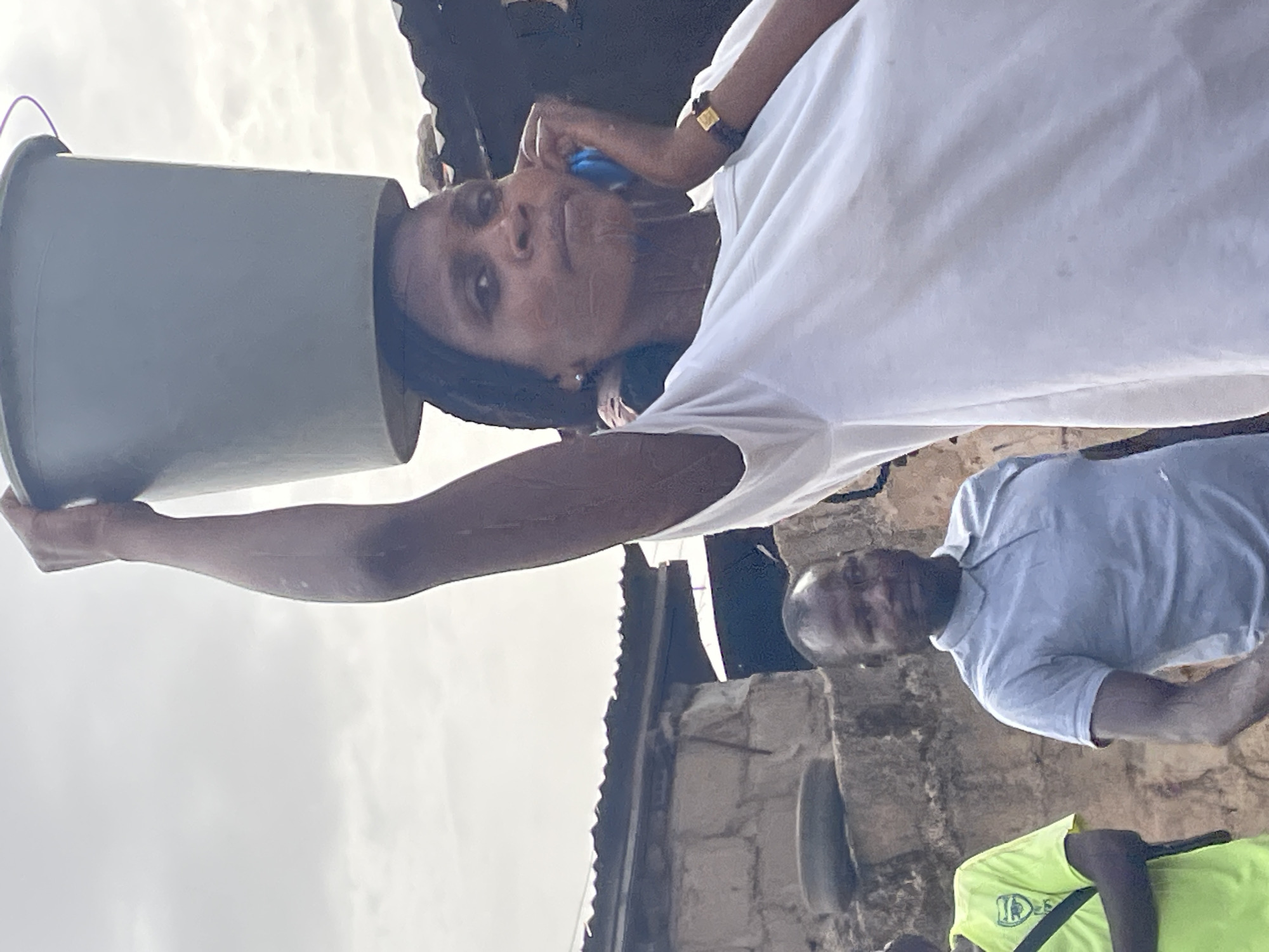 In an outdoor setting in Jos, Nigeria, a dark-skinned woman in a white t-shirt holds a gray bucket aloft. The bucket is angled, as if pouring its contents. The woman is facing the camera, with a blue object near her ear. An additional man in a light blue shirt is visible in the background. A building structure with a corrugated roof is also in the scene. Other individuals are partially visible in the scene.