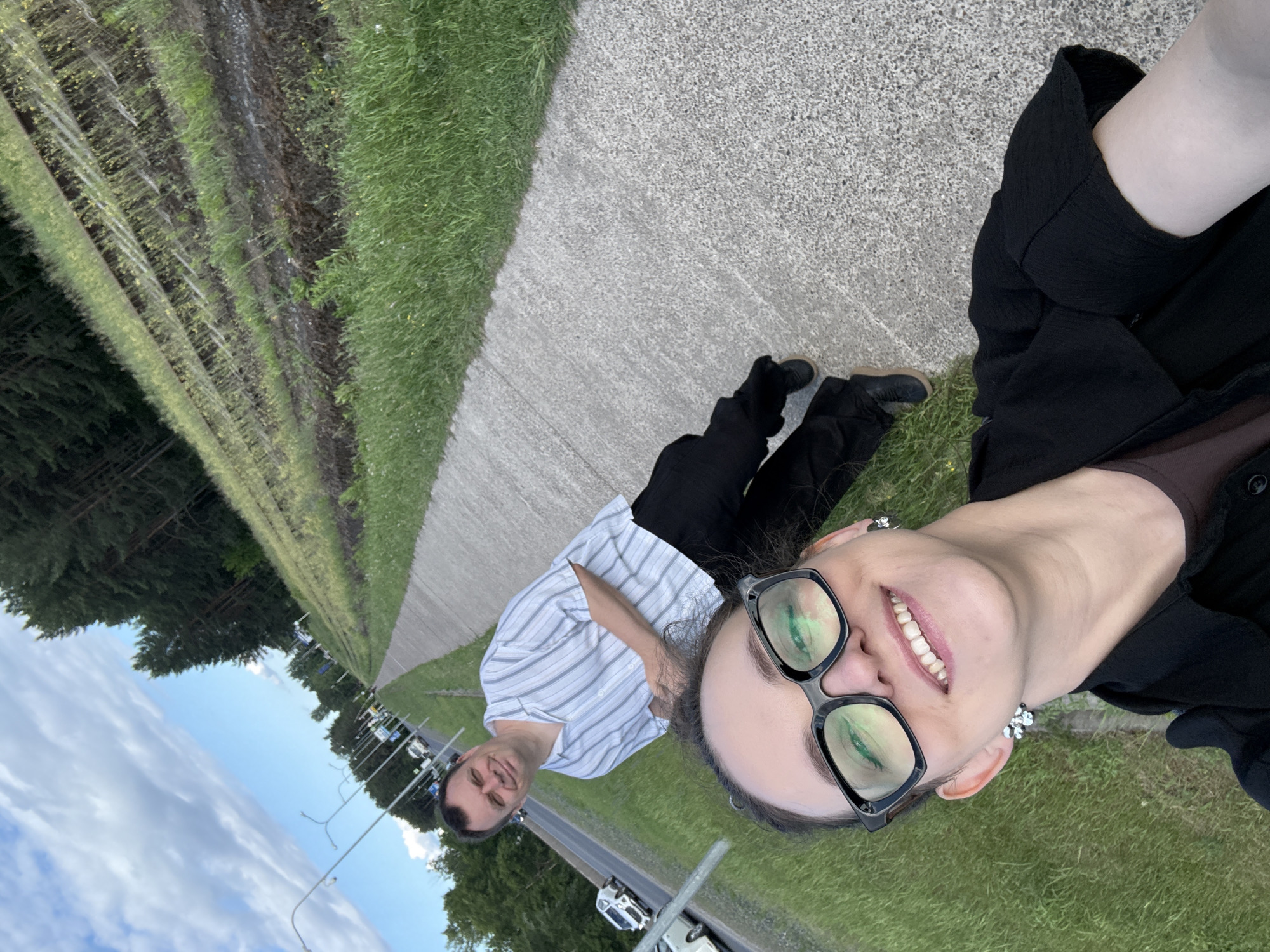 This is a selfie taken outdoors in Chaikovsky, Russia. In the foreground, a woman with dark hair and glasses smiles at the camera. She is wearing a black shirt. Behind her, a man in a striped shirt and black pants stands on a paved walkway. To the left of the walkway is a road with cars, and to the right is a grassy embankment with a line of trees in the distance. The sky is partly cloudy.