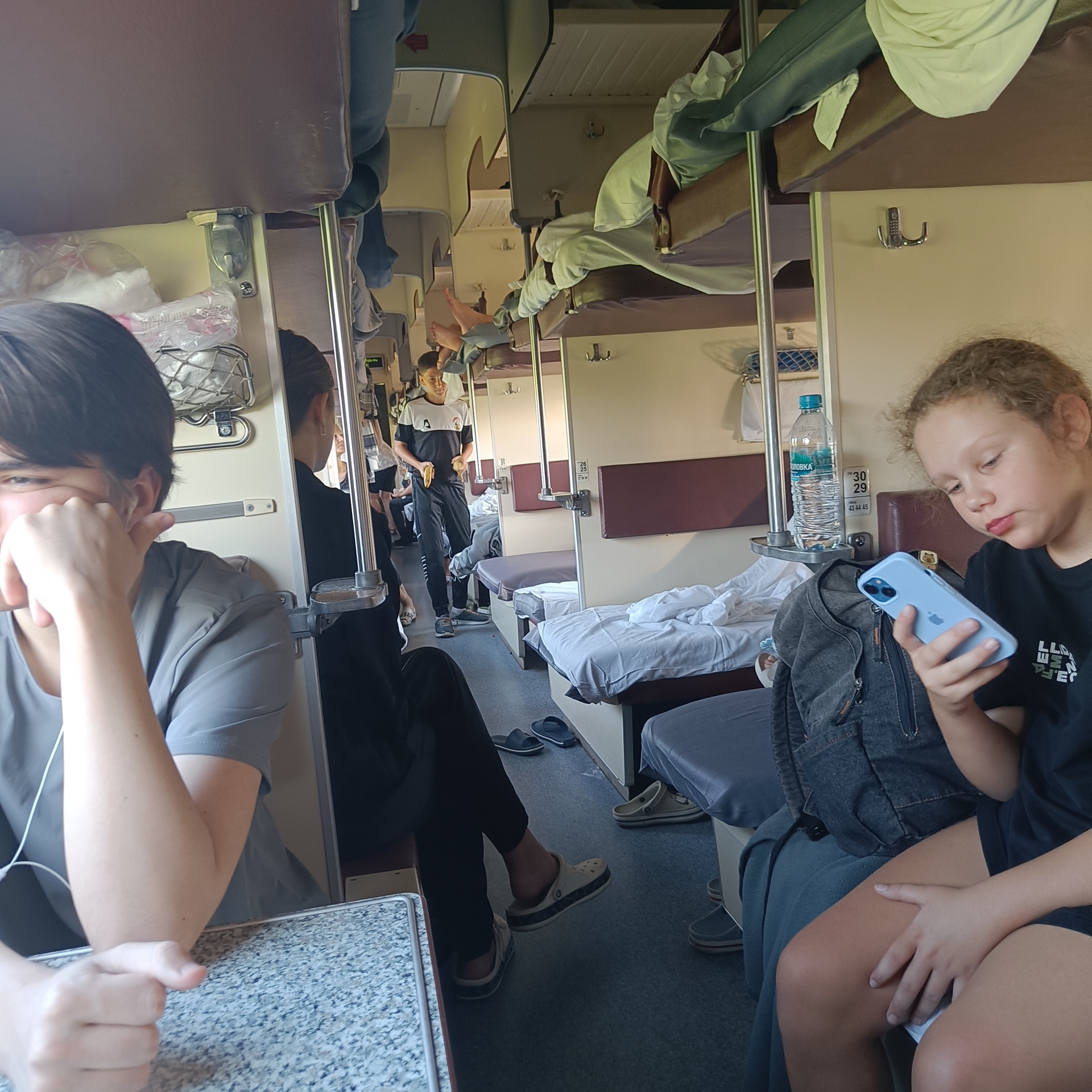 The image depicts the interior of a train compartment, likely a sleeping carriage, with multiple bunks visible. In the foreground, on the left, a person with dark hair is seated, resting their head on their hand, with headphones visible. To their right, another person is partially visible. Further down the compartment, a young boy in a dark and white t-shirt and black pants stands, holding a yellow object. On the right side of the image, a young girl with curly hair is seated, looking at a light blue smartphone. Several other individuals and their legs are visible on the bunks and the floor, suggesting a crowded environment. Bags and personal belongings are scattered around. The environment appears utilitarian, typical of train travel, with beige walls and brown upholstery on the bunks. A bottle of water is placed on a shelf. The overall scene captures a moment of transit within a train. There is no visual information in the image to suggest the specific location within the city of Mineralnye Vody, Russia.
