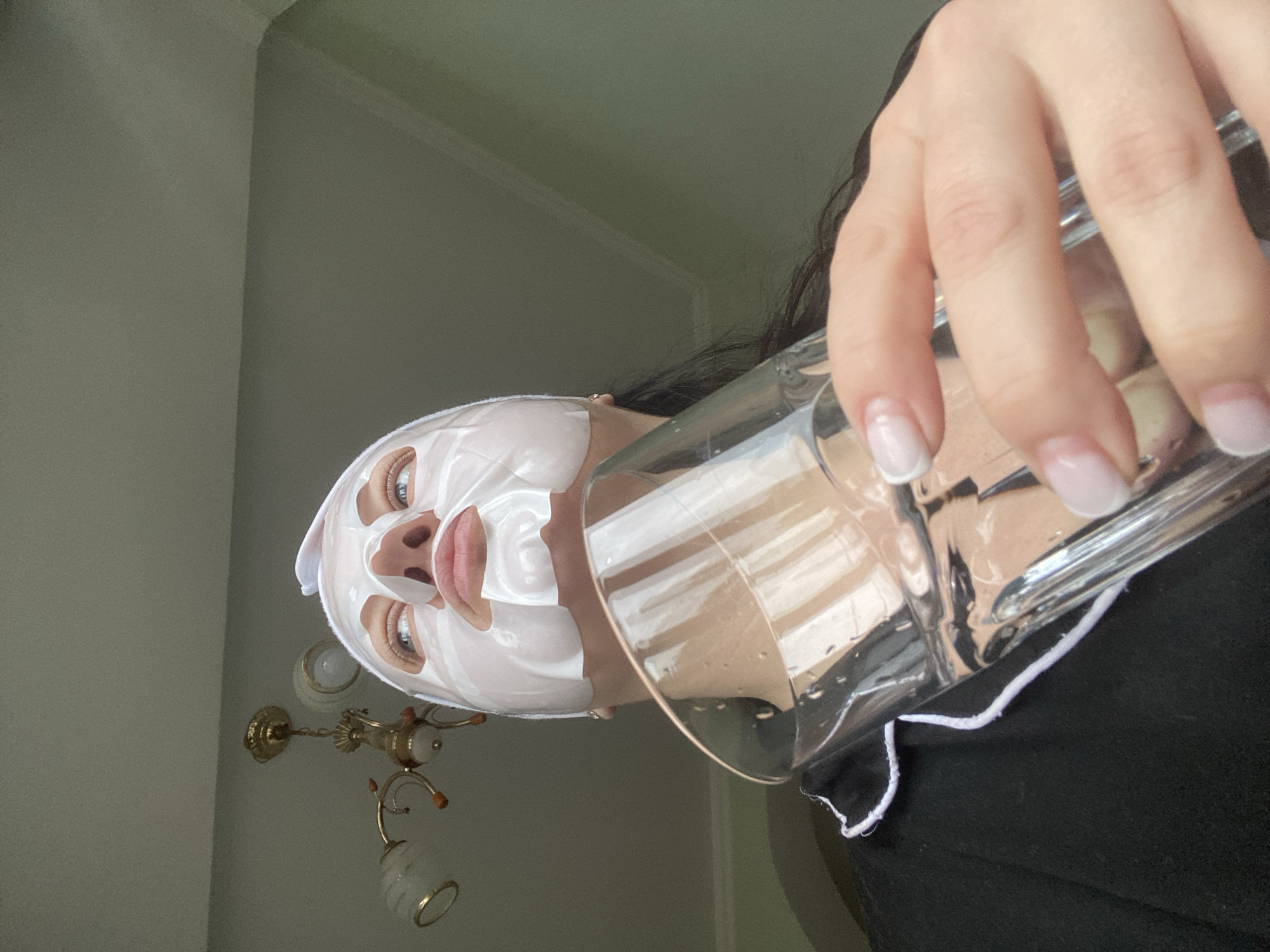 A person wearing a white sheet mask is holding a clear glass in their right hand. The person's face is slightly tilted and is positioned in the upper left quadrant of the image. The glass is angled towards the viewer, and the person's hand is partially obscuring the glass. The background features a light green wall, and a chandelier is visible in the lower left corner. The person is wearing a black garment with white trim. There is no indication of this photo's location being in Sochi, Russia.