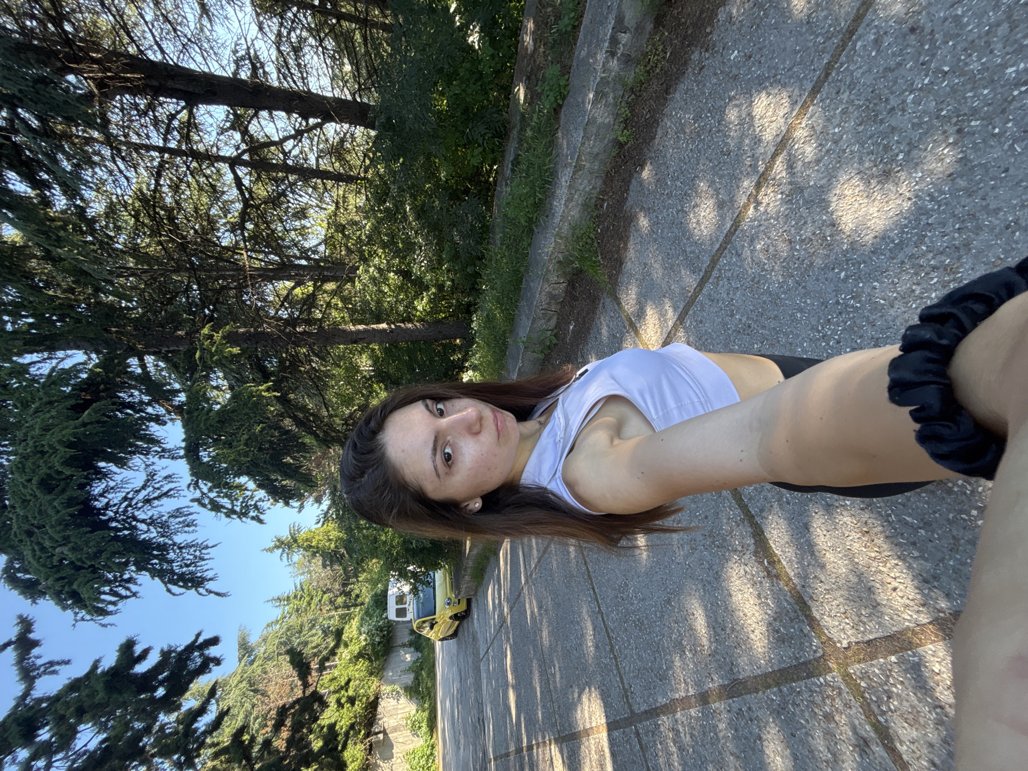 A young woman with long dark hair stands on a paved path outdoors, taking a selfie. She is wearing a white athletic top and dark bottoms. Behind her, a yellow car and a white van are parked on the side of the road, which is lined with trees. The scene appears to be in a park or a scenic area, possibly in Varna, Bulgaria, given the presence of Mediterranean flora and the architectural style of the wall on the left. The sun is casting dappled shadows on the path.
