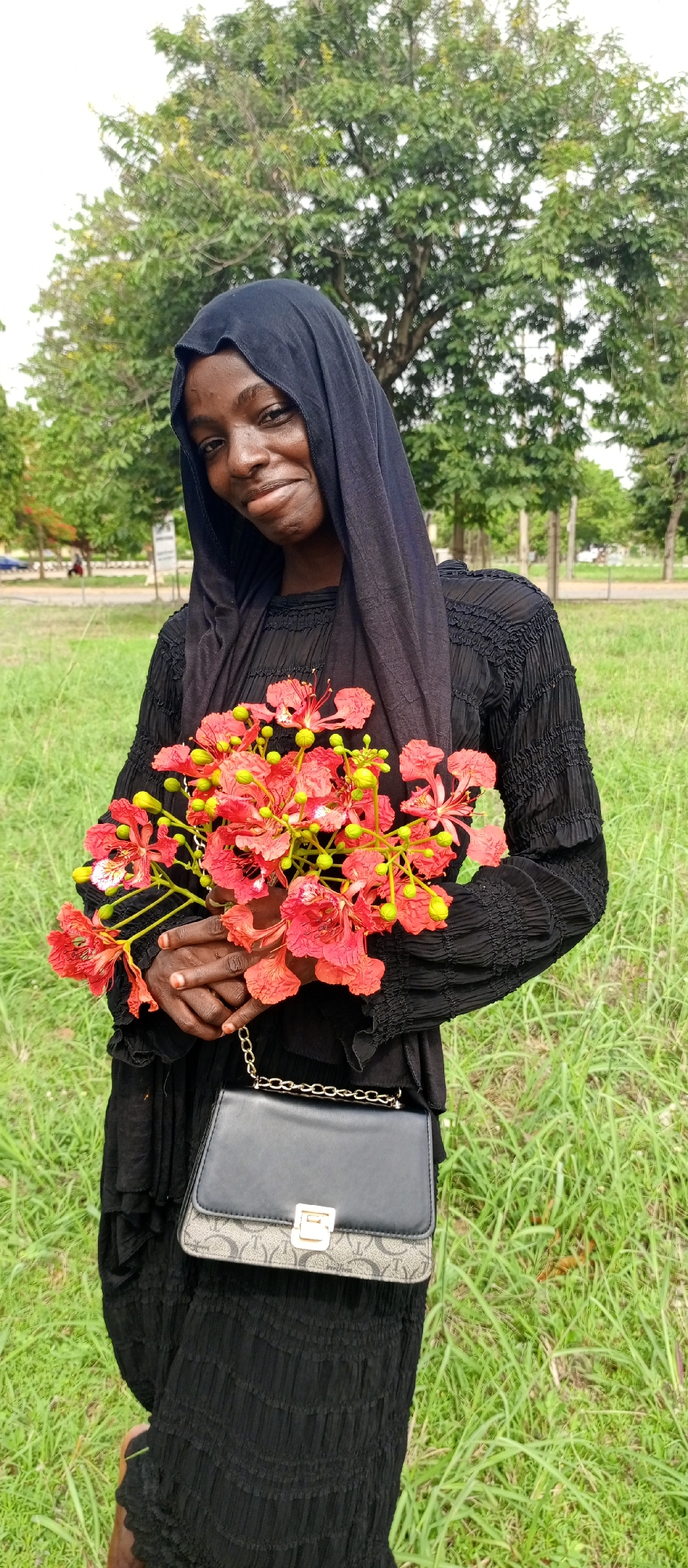 A young Black woman stands outdoors, smiling gently at the camera, holding a vibrant bouquet of red and green flowers. She is dressed in a modest black long-sleeved dress and a matching black hijab that frames her face. Her dark skin contrasts with the bright red petals and green buds of the flowers, which appear to be from a Flamboyant tree (Delonix regia). Her hands are clasped around the flower stems.
Across her body, a black and grey patterned purse with a golden chain strap and a matching gold-toned clasp is visible. The setting is a lush green outdoor area, likely a park or field in Keffi, Nigeria, as indicated by the location context. In the background, verdant trees with dense foliage are visible under a bright sky, suggesting daytime. The ground is covered in green grass, with some areas appearing more manicured than others. The woman's expression is one of calm happiness and warmth. There is no visible text in the image.
