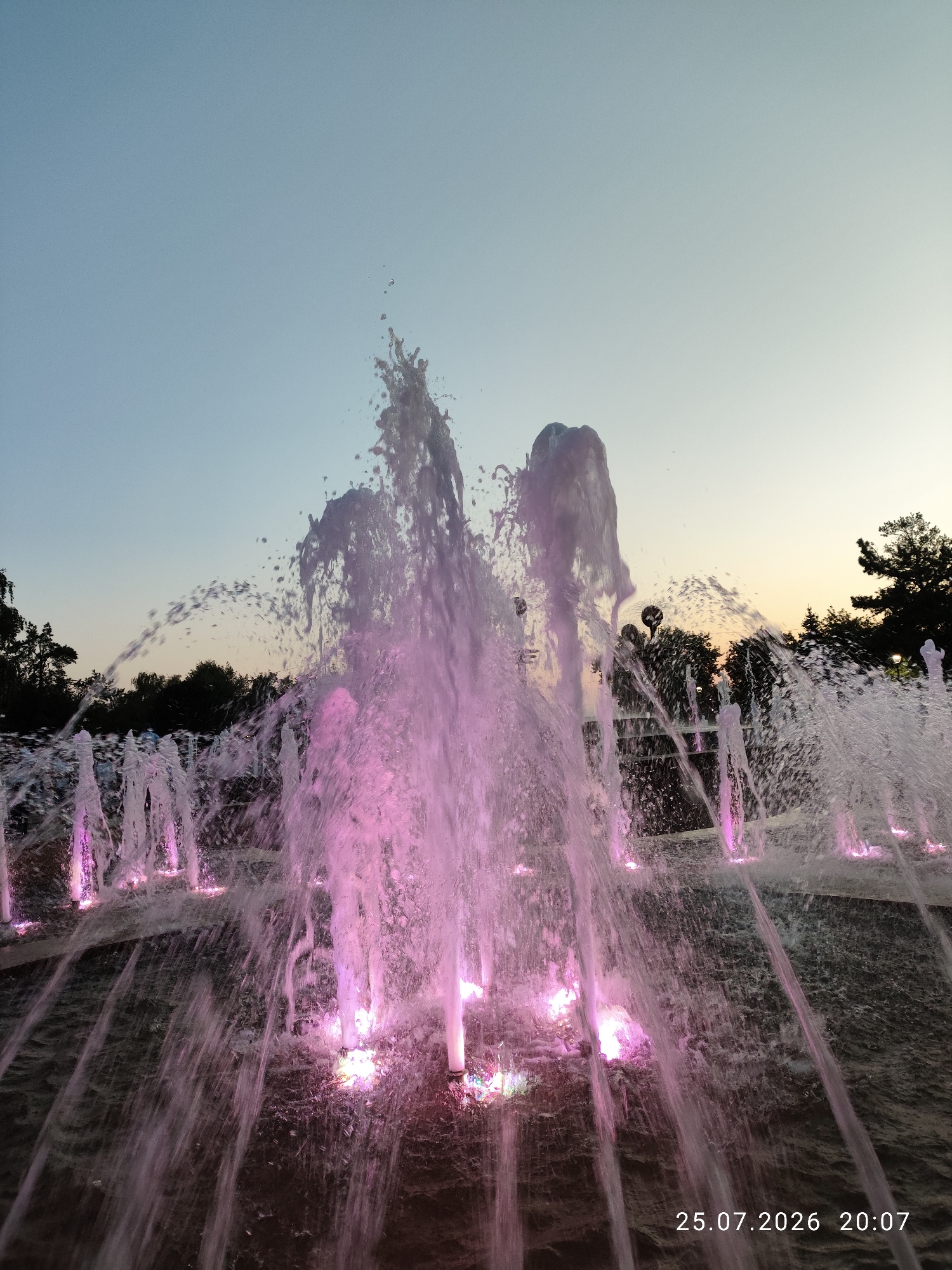 The image shows a fountain with illuminated water jets in the city of Pavlodar, Kazakhstan. The fountain is lit with pink and purple lights, creating a vibrant display. The water is spraying upwards and outwards, with many droplets visible. In the background, there are trees and a clear sky, suggesting it is either dusk or dawn. A timestamp in the bottom right corner reads "25.07.2026 20:07", indicating the date and time the photo was taken. There are no people visible in the image.