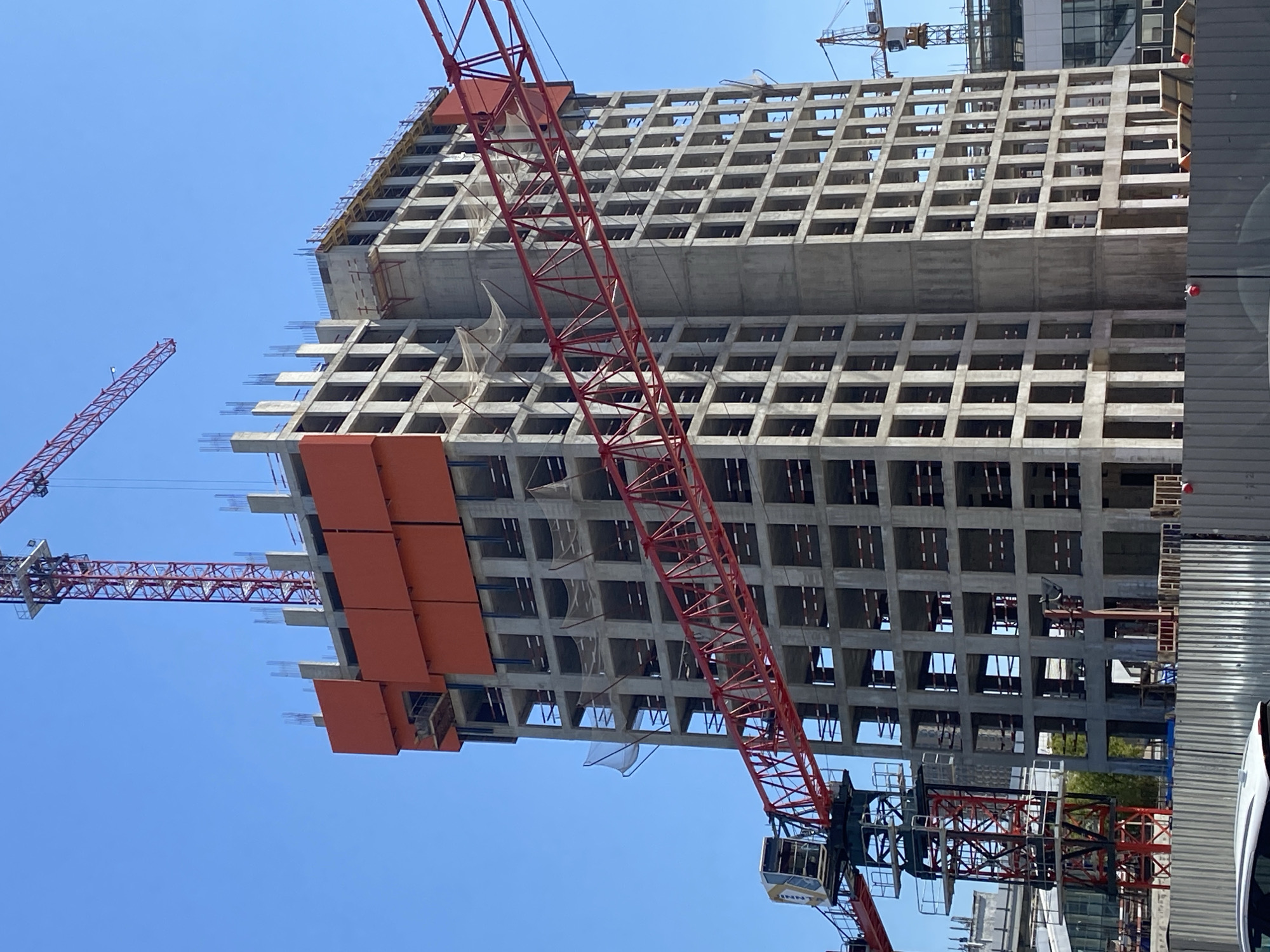 An outdoor, eye-level, vertical medium shot captures a high-rise building under construction in Moscow, Russia, under a clear blue sky. The building is a grey concrete structure with exposed floors and openings. Orange panels are affixed to the upper section of the building. Two red construction cranes are visible, one in the foreground, its boom extending across the frame, and another taller crane positioned behind and to the right of the main building. In the background, other buildings and construction cranes can be seen. A grey metal fence runs along the bottom of the image, with a portion of a white car visible in the lower left corner. No people are discernible in the image.