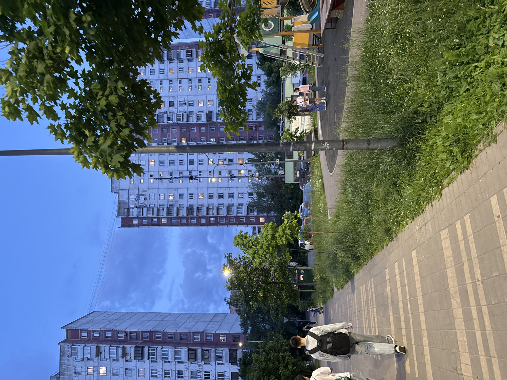 The image depicts an urban residential scene during dusk or early evening. Large, multi-story apartment buildings, characterized by their numerous windows with many showing interior lights, dominate the background. Their facades exhibit a mix of light-colored walls and darker, possibly brick-colored, sections.
In the foreground, a paved sidewalk stretches diagonally across the lower half of the frame, lined on one side by a strip of lush green grass dotted with small white wildflowers. A lamppost stands tall beside the sidewalk. Several individuals are present: a person wearing a light jacket and a black backpack walks away from the camera in the foreground, while a small group of three people stands further down the sidewalk near a colorful playground structure that includes a slide and climbing elements.
Trees with dense green foliage are prominent, with a large leafy branch extending into the upper left portion of the scene. The sky is a deep blue, indicative of twilight, with some clouds visible between the distant buildings. Faint, parallel shadows are cast across the sidewalk, suggesting light coming from an external source or a structure above. Cars are parked along a street visible in the mid-ground, behind the playground and trees. No discernible text is clearly visible in the image.