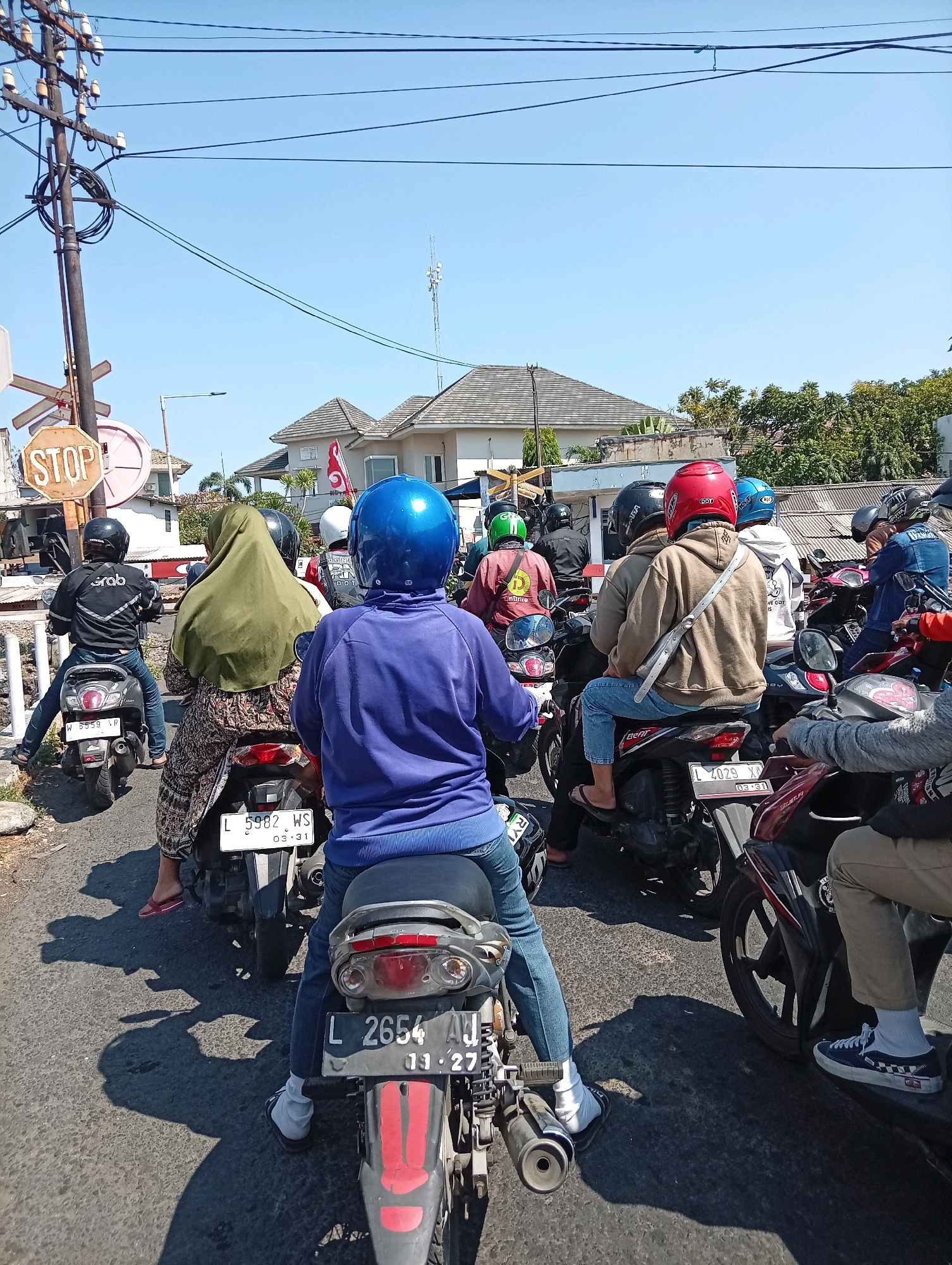 This is a medium-angle, eye-level shot of a traffic jam with numerous motorcycles stopped on a paved road. The scene is set in Surabaya, Indonesia, under a clear blue sky. In the foreground, the rear of a motorcycle with a black license plate labeled "L 2654 AJ" is visible. A person wearing blue jeans and white socks is seated on this motorcycle. To the left, another motorcycle with a black license plate "W 5559 VP" is stopped, with a person in blue jeans and black sandals seated on it. A woman wearing a green hijab and a patterned dress is riding on the back of a motorcycle to the left of the center. Further back, a rider in a brown hoodie and red helmet is on a motorcycle, and to their right, a person in a blue hoodie and blue helmet is also on a motorcycle. Other motorcycles and riders are visible in the midground and background, indicating a significant traffic backup. A prominent "STOP" sign on a pole is located to the left of the frame, along with other traffic signals. Buildings with tiled roofs and trees are present in the background. The sunlight casts long shadows on the road, suggesting it is daytime.
