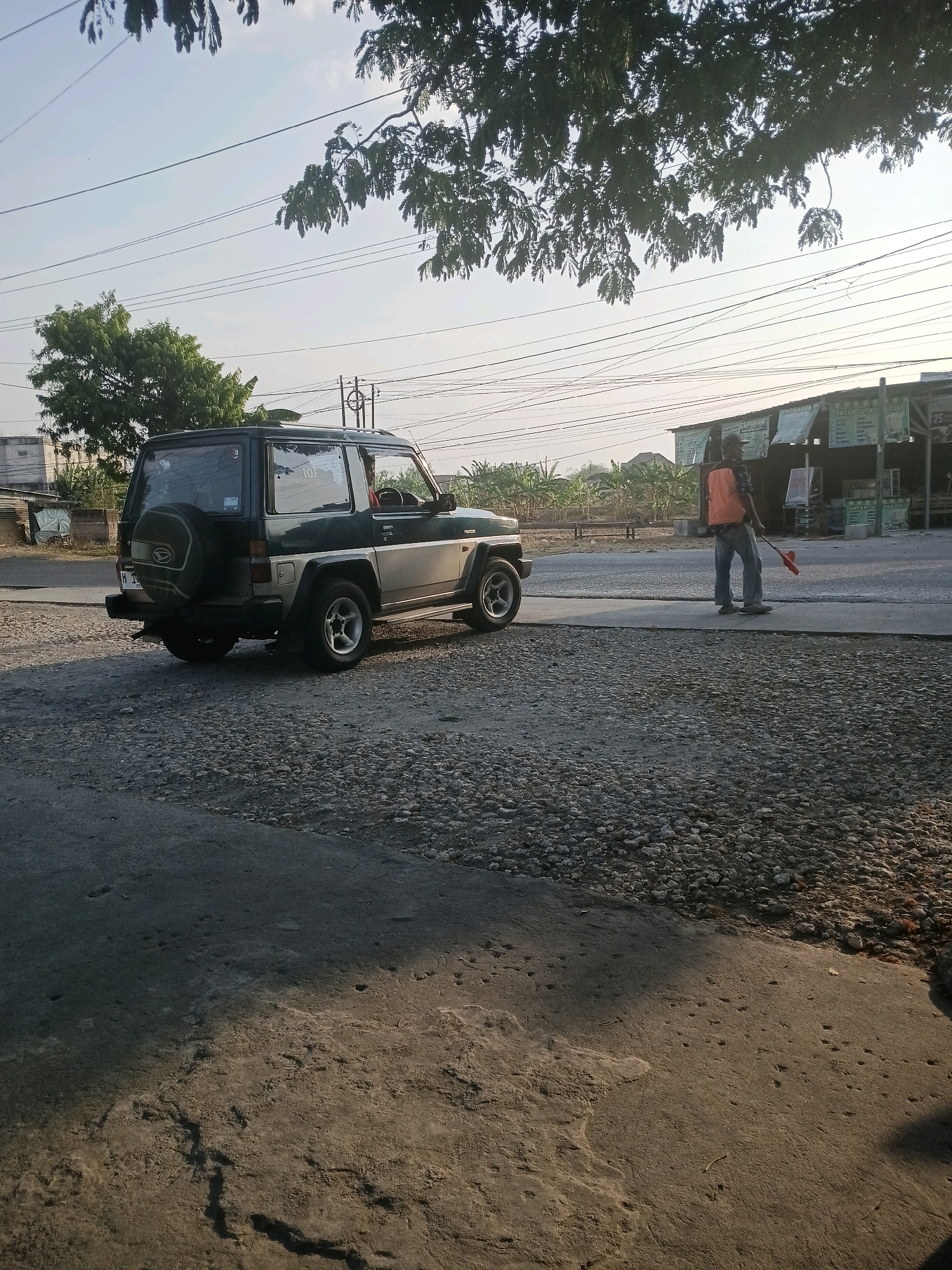 The image captures a scene in Purwodadi, Indonesia, under a clear sky with dappled sunlight filtering through tree branches overhead. In the foreground, a dark green and beige Daihatsu Rocky SUV is parked on a gravel and concrete surface. The vehicle has a spare tire mounted on the rear. To the right of the SUV, a person wearing a bright orange vest, blue jeans, and dark shoes is standing on the paved road. They are holding a red object, possibly a broom or a flag, and appear to be performing some task. Behind the person, a row of buildings with signs, including one that reads "IZEN JUSSU TASU", line the street, and beyond them, a field of banana trees is visible. The environment suggests a roadside setting in a rural or semi-urban area.