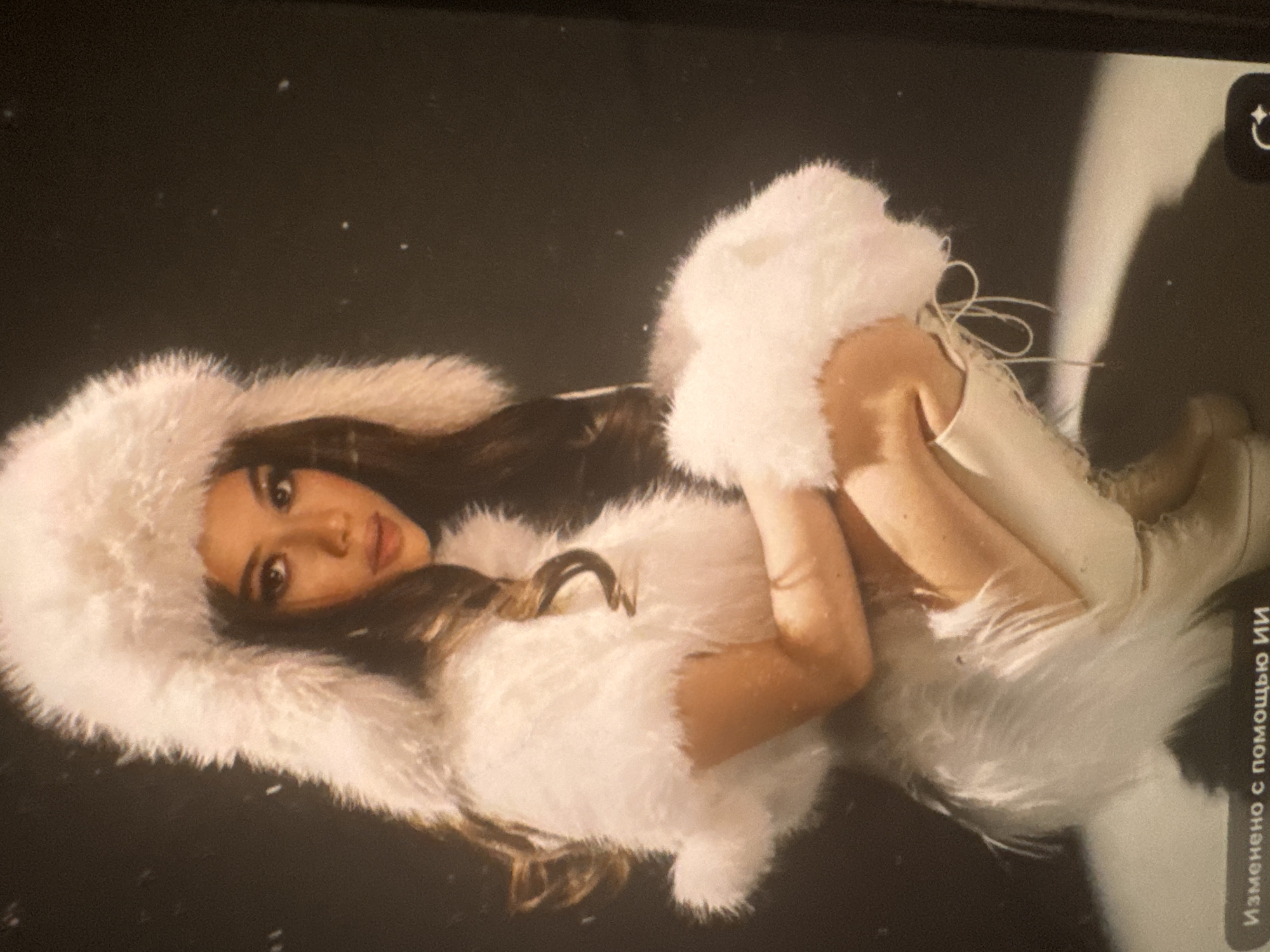 The primary image is a photograph of a screen displaying another image, as evidenced by screen reflections and visible interface elements. The central subject is a young woman, likely in her late teens or early twenties, with dark hair and a serene expression, looking towards the viewer. She is posed reclining or lying down, dressed in a distinctive all-white outfit featuring extensive faux fur trim on a hood, a vest or top, and knee-high lace-up boots. Her body is curved, with her arms bent and one hand gently resting near her face.
The setting appears to be a studio or a controlled environment, characterized by a dark, possibly black, background. Small, light specks are scattered across this dark backdrop, suggesting either dust, ambient light reflections, or stylized elements resembling falling snow. The overall aesthetic hints at a winter, fantasy, or glamorous theme.
Notable details include the texture of the plush white fur, the intricate lacing on her boots, and her defined facial features. Visible in the top right corner of the screen reflection is a white camera-related icon, and along the bottom right edge, Russian text reads "Изменено с помощью ИИ," which translates to "Changed with the help of AI." This text indicates that the image displayed on the screen has undergone some form of artificial intelligence-assisted modification. The image is presented rotated 90 degrees counter-clockwise.