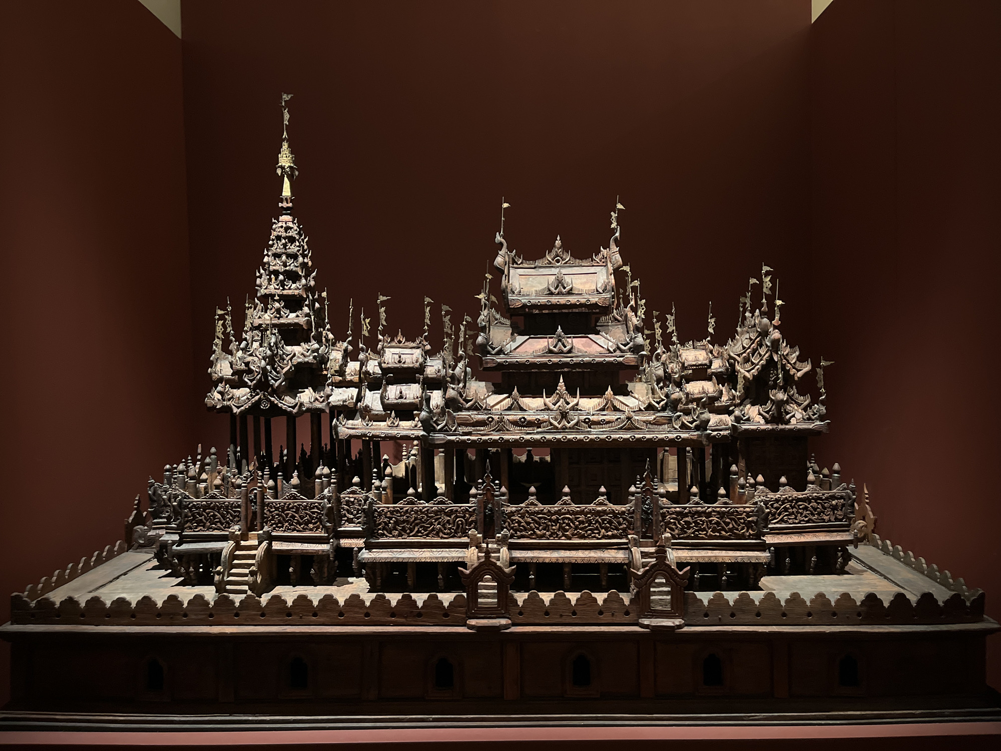 The image shows a detailed, ornate wooden model of a temple or palace, likely of Southeast Asian origin. The model is displayed against a dark, reddish-brown background, possibly a museum wall. The structure features multiple tiered roofs with intricate carvings, spires, and decorative elements. Small flags adorn the peaks of some structures. The base of the model is also wooden and appears to have decorative panels and small arched openings. The lighting highlights the detailed craftsmanship and dark wood of the model. There are no people visible in the image. The scene is static and presented as an exhibit. There is no information to suggest this model is located in Paris, France.