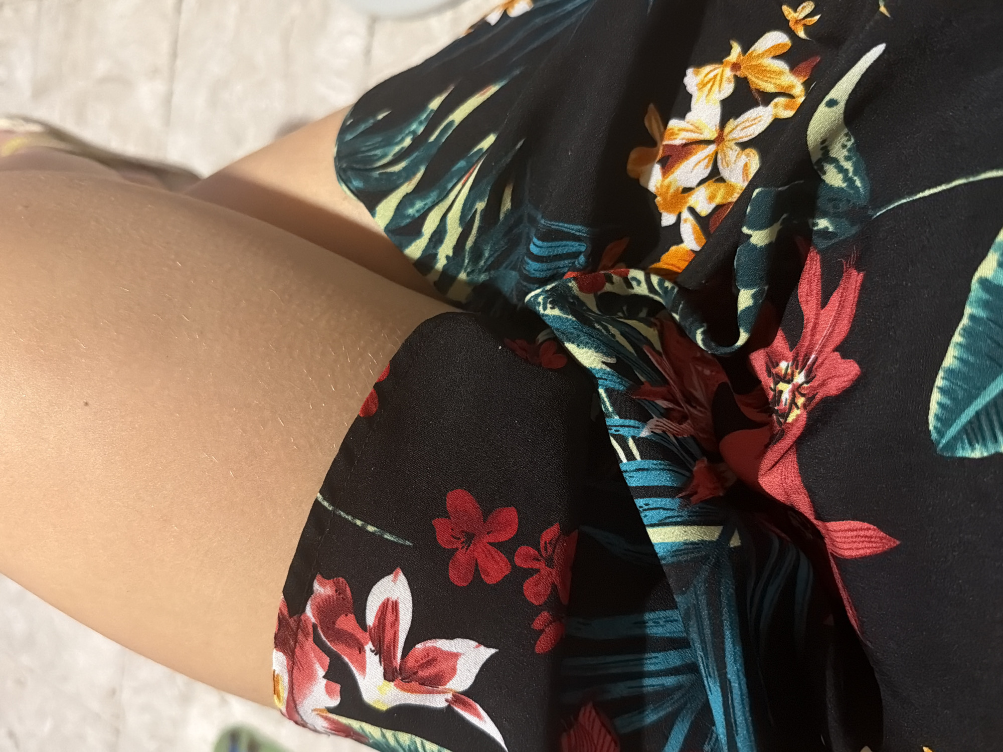 The image shows a close-up view of a person's legs, crossed at the knees, wearing a black dress with a tropical floral print. The dress features large red and white flowers, as well as smaller red flowers, and stylized green and yellow leaves. The background is out of focus and appears to be a light-colored, textured surface, possibly a floor or wall. The lighting suggests an indoor setting with soft, ambient light. There is no discernible location in the city of Kiti, Cyprus, depicted in the image.
