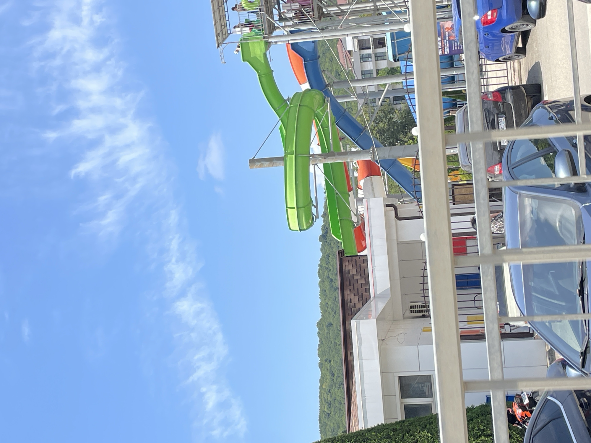 The image shows a water park with slides in Dzhubga, Russia. The slides are brightly colored, with green, blue, and orange sections. There are people visible on the platforms and slides of the water park. In the foreground, there are cars parked in a lot, with a white fence separating the parking area from the water park. The sky is blue with some wispy clouds. A lush green forested hill is visible in the background.