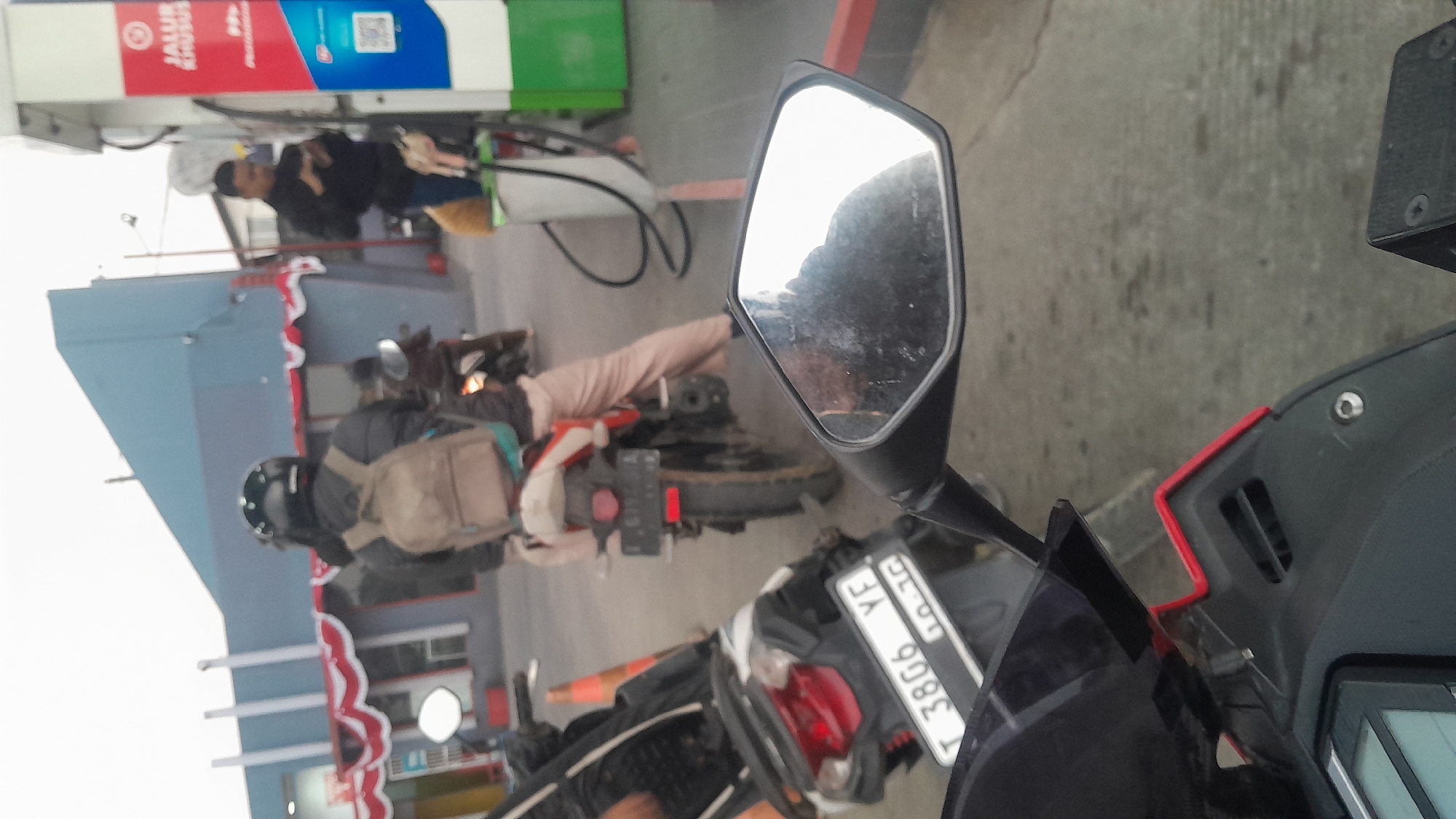 At a gas station in Subang, Indonesia, several individuals on motorcycles are waiting in line. A person wearing a black helmet and a backpack is seated on a motorcycle. Another person in a black jacket is standing at a gas pump, looking at their phone. In the foreground, a motorcycle's rearview mirror reflects the sky and a portion of the gas station. The gas station has a blue and white building with red and white decorative banners.