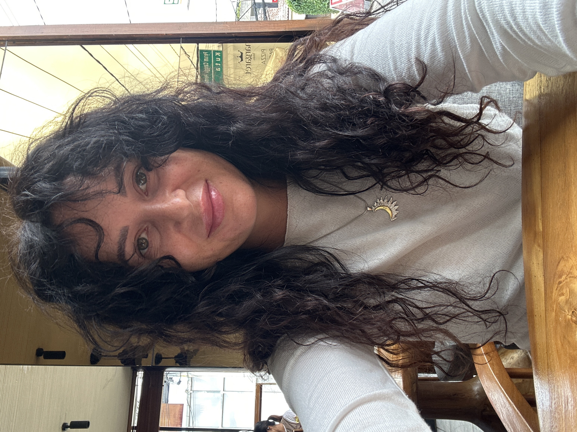 A woman with long, dark, curly hair is taking a selfie in what appears to be a cafe or restaurant in North Kuta, Indonesia. She is wearing a white long-sleeved shirt and a silver necklace with a sun and crescent moon pendant. She is seated at a wooden table, and her hair falls over her shoulders and onto the table. In the background, there are reflections of lights and what appears to be a street scene through a window. A sign with Indonesian text is visible in the background.