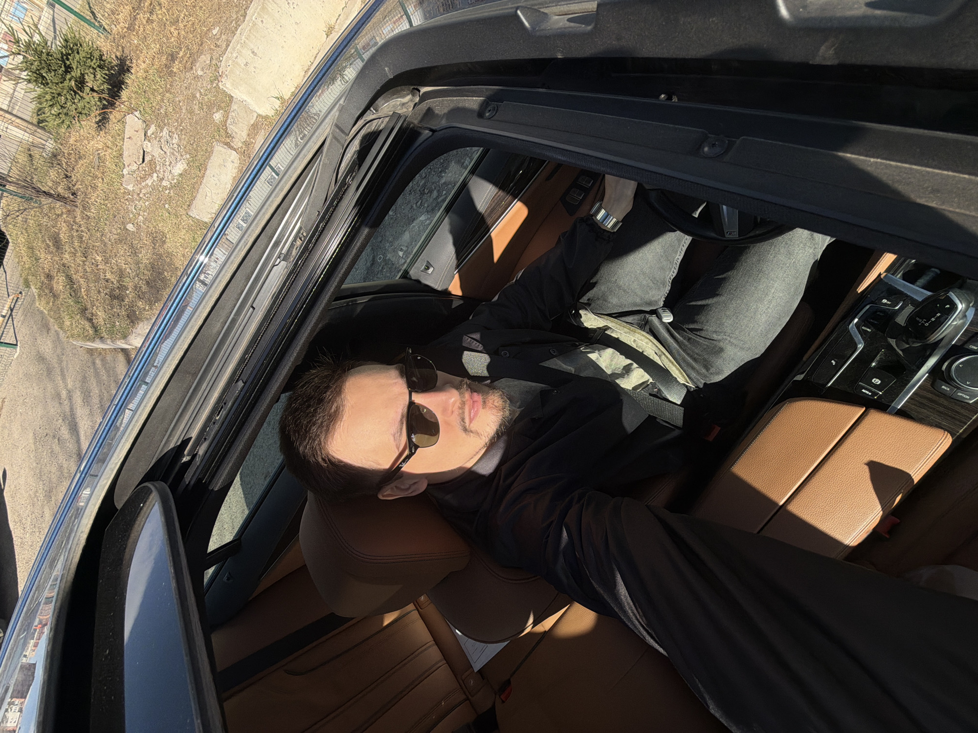 The image presents a man relaxing inside a car, viewed from an overhead perspective through an open panoramic sunroof. The subject is a man with a short beard and dark hair, wearing sunglasses, a black long-sleeved top, and grey jeans. He is leaning back against a brown leather headrest, with strong sunlight illuminating parts of his face and the car's interior, suggesting a bright, sunny day, likely midday or early afternoon. His right arm is extended, indicating he may be taking a selfie.
The car's interior features luxurious brown leather upholstery on the seats and center console. A black dashboard and steering wheel are visible, with an "M" logo clearly seen on the steering wheel, suggesting a BMW M Sport model or similar. Buttons on the center console are labeled "AUTO H" and "ECO PRO".
Outside the car, a patch of ground with dry grass, dirt, and some concrete slabs is visible, along with a small green bush or tree and a metal fence in the background. The scene depicts a vehicle parked outdoors next to a somewhat unkempt, natural area. The location context for this scene is Krasnoyarsk, Russia.