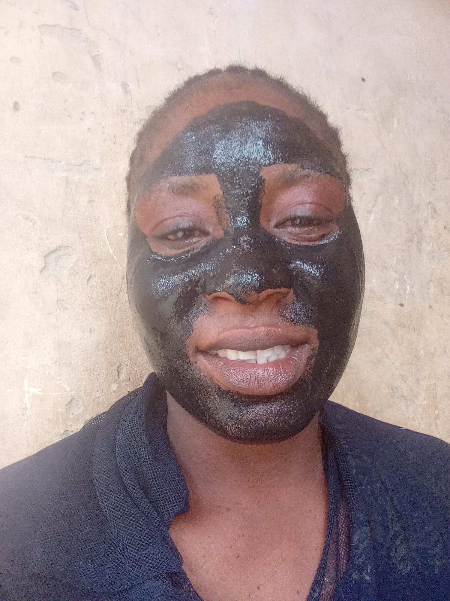 A close-up, front-facing portrait features an adult individual, likely female, with dark skin, wearing a black facial mask. The mask is applied to most of the face, including the forehead, cheeks, nose, and chin, but leaves the eyes, eyebrows, and mouth exposed. The mask appears somewhat glossy or wet and is applied with some visible texture. The individual looks directly at the camera with a slight smile, revealing their upper teeth, and their eyes are open. Short, dark hair is visible around the hairline.
The subject is wearing a dark blue garment, possibly a top or dress, made of a mesh-like or lace fabric, visible on the shoulders and chest area. The background is a plain, light-colored wall with a rough, weathered texture, suggesting stucco or plaster. The lighting is bright and natural, indicating a daytime setting. No text is visible. The scene is located in Akwanga, Nigeria.