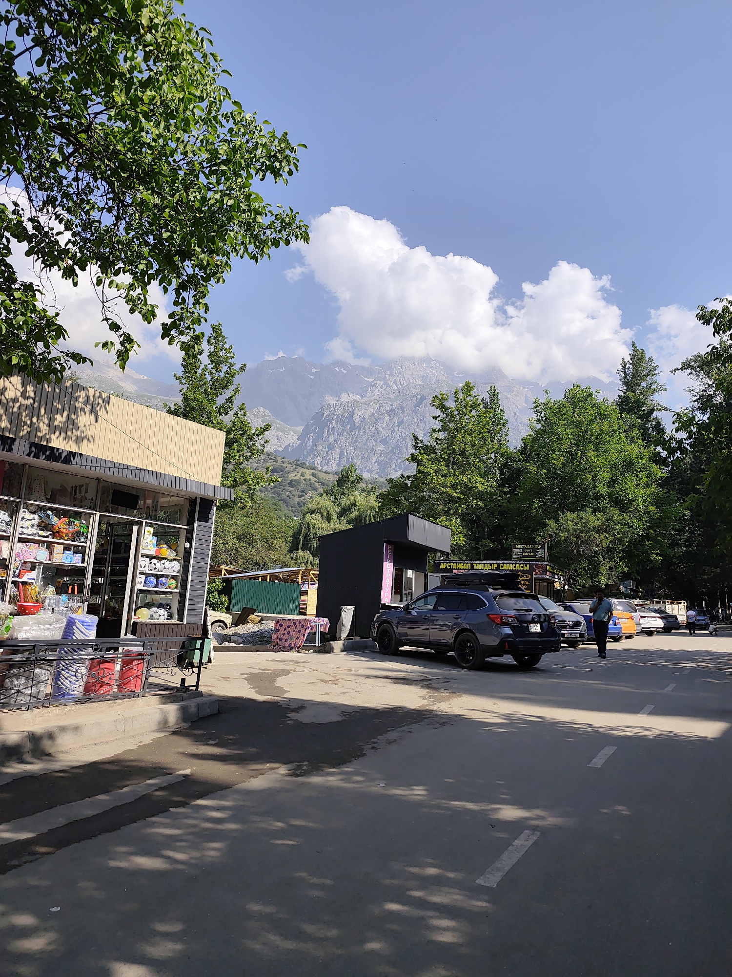 This image depicts a street scene in Arstanbap, Kyrgyzstan, with a clear blue sky and fluffy white clouds dominating the upper portion. In the foreground, a paved road with white lane markings is visible, partially shaded by the leaves of trees. To the left, a small, modern-looking shop with a glass storefront displays various items, including what appear to be toys and household goods. Adjacent to the shop, a black building with a dark roof stands. Further down the road, several cars are parked, including a dark gray SUV in the foreground. A person in a light blue shirt is walking along the sidewalk near the parked cars. In the background, lush green trees line the street, leading up to a majestic, rocky mountain range under a bright sky. The overall scene suggests a sunny day in a town nestled against a mountainous backdrop.