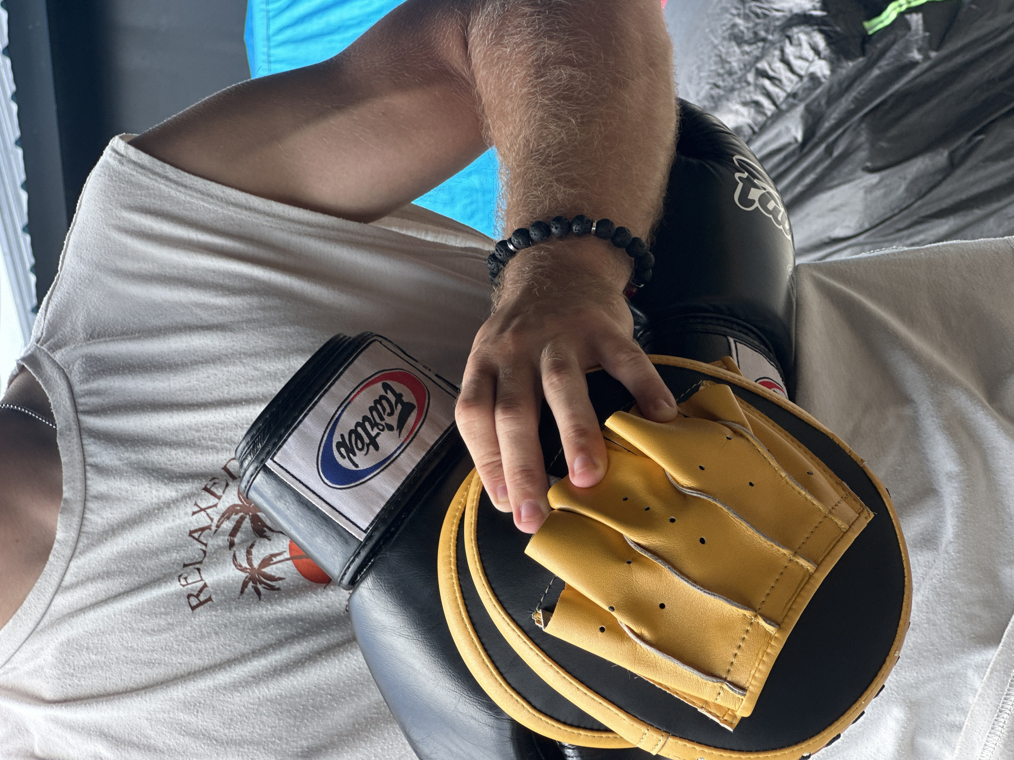 A person, with visible arm hair and wearing a white sleeveless t-shirt with a "RELAXED" palm tree graphic, holds a pair of black and yellow boxing mitts. The mitts have a "Fairtex" logo on them. The person is wearing a dark beaded bracelet on their left wrist. The background includes a grey surface and a bright blue tarp. The scene appears to be outdoors, possibly in a training facility in Chalong, Thailand.