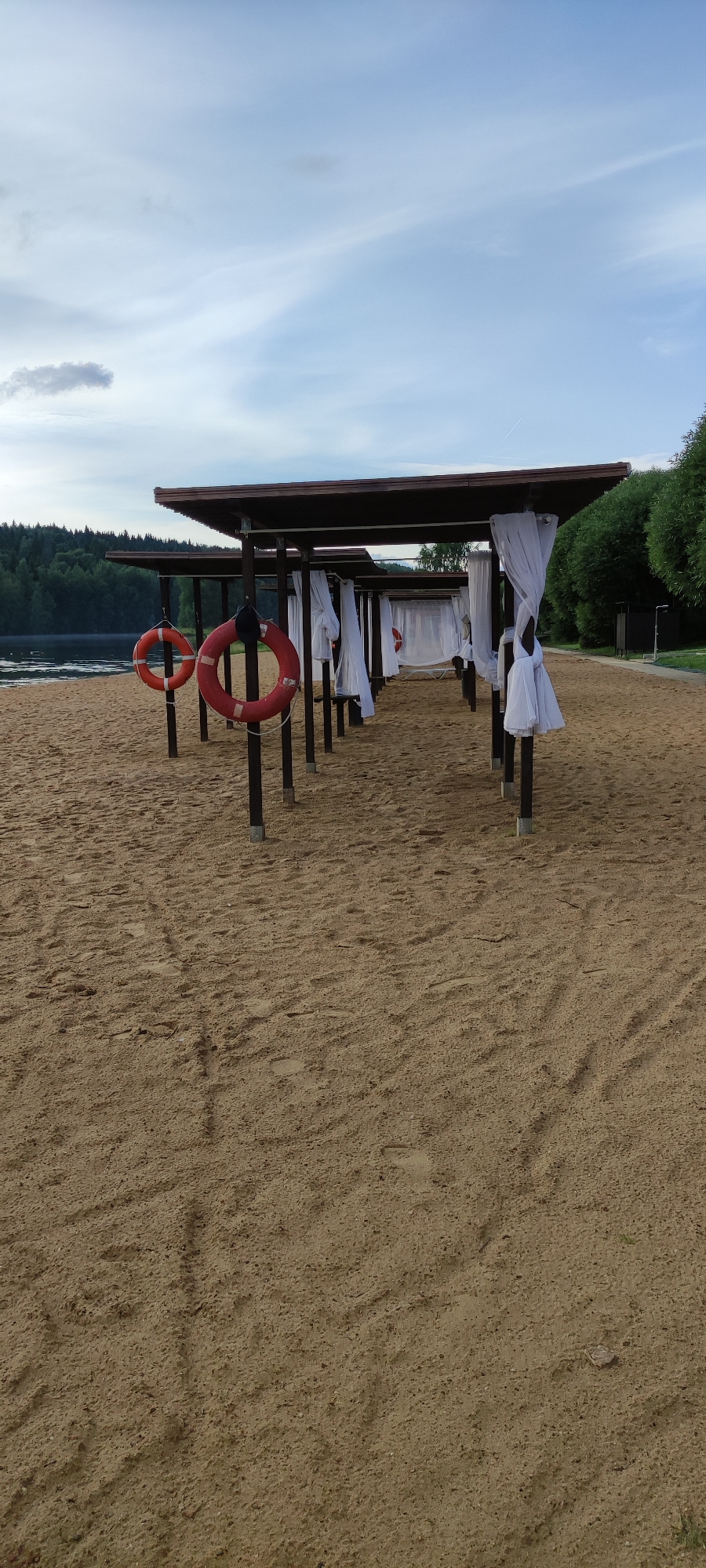 This media file captures a serene beach scene with a row of cabanas lined up along the sand. The cabanas are covered with white drapery, creating a sense of privacy and relaxation. Two bright red life preservers hang prominently on poles in the foreground, suggesting a proximity to water and a focus on safety.
The beach is made of light-colored sand, marked with footprints, indicating recent activity. In the background, a calm body of water is visible, bordered by a dense forest of green trees under a partly cloudy sky. The time of day appears to be late afternoon or early evening, with soft lighting and a tranquil atmosphere.
There are no people visible in the image, giving it a peaceful and uncrowded feel. The weather seems mild and pleasant, conducive to outdoor activities. The overall impression is one of a well-maintained recreational area, possibly a resort or a public beach, designed for leisure and relaxation by the water. The presence of life preservers and the forested backdrop are key location cues. No text is visible in the image.