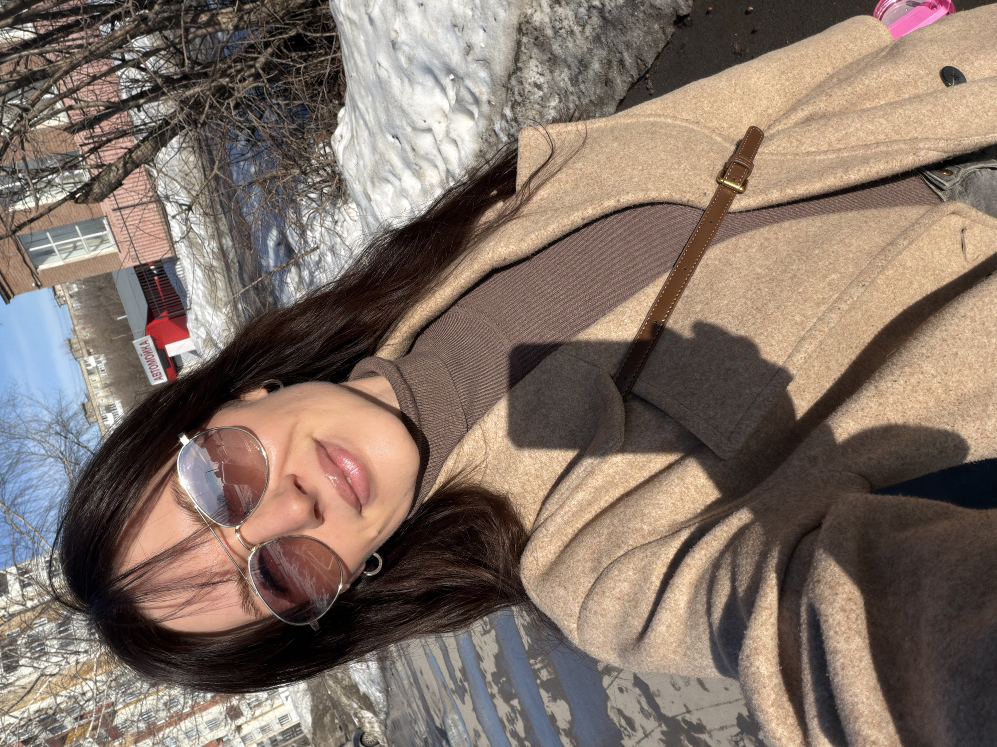 This image is a close-up, selfie-style photograph of a female individual in an outdoor urban setting in Perm, Russia, identifiable by a prominent "АВТОМОЙКА" (CAR WASH) sign in the background. The individual has dark, shoulder-length hair with bangs and wears reflective aviator-style sunglasses with pinkish lenses. Her lips are glossy, and she displays a slight smile. She is adorned with small silver hoop earrings. Her attire consists of a brown ribbed turtleneck sweater underneath a tan wool-blend coat. A brown strap with a brass buckle crosses her upper torso. A partial pink object is visible on the lower right of the frame. The scene is illuminated by strong sunlight, casting distinct shadows, and patches of melting snow are present on the ground, indicative of a late winter or early spring season. The background includes a multi-story, light-colored residential building to the left, and a red-brick building beyond a cluster of bare trees and bushes to the right, adjacent to the identified car wash facility with a red barrier. A clear blue sky is visible above the urban landscape. The individual is actively taking the self-portrait; no other persons or interactions are depicted.