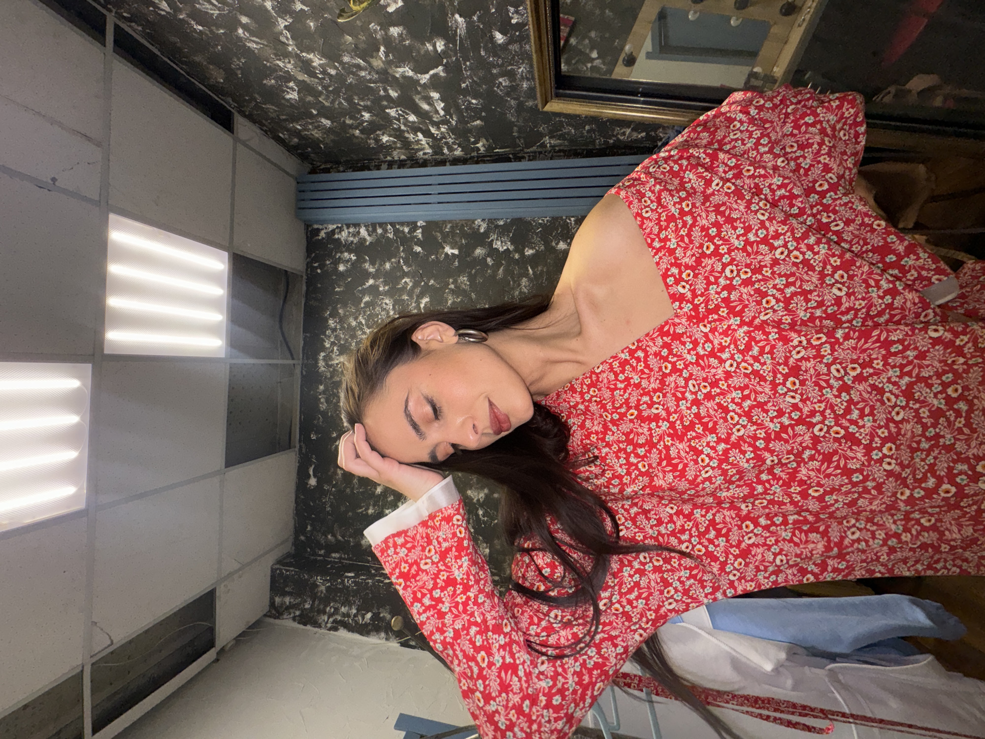 A woman with long dark hair wearing a red floral dress with white cuffs and sleeves, and a silver hoop earring is resting her head on her hand. The room has two different wall designs: one side features a white, grid-like panel with illuminated square sections, and the other has a black textured finish with blue piping. There is a framed mirror on the textured wall. Other objects are visible, including clothing on a metal rack and possibly other furniture. I am unable to tell the location within the city of Krasnoyarsk based on the image.