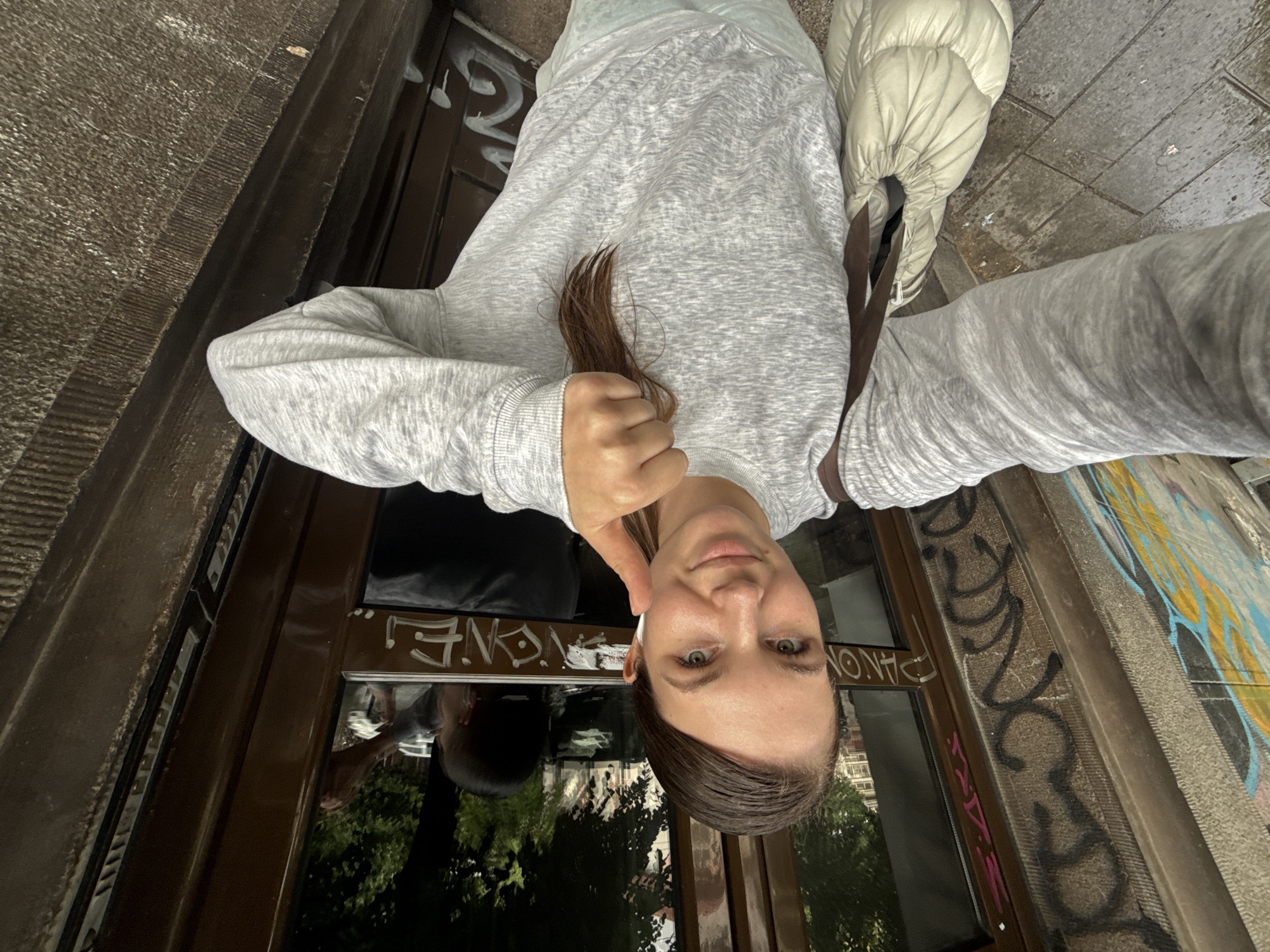 A selfie shows a woman with green eyes and brown hair in front of a storefront in Belgrade, Serbia. She is wearing a light gray sweatshirt and a white puffer jacket over her shoulder. She is holding a strand of her hair. The storefront window reflects trees and buildings. Graffiti is visible on the wall beside the storefront.