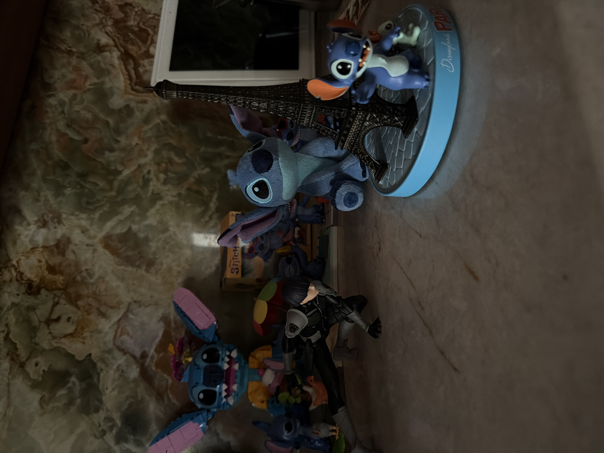 A collection of Stitch figurines and a character from the anime "Fullmetal Alchemist" are displayed on a marble surface in front of a marble wall. In the foreground, to the right, a small Stitch figurine stands on a blue base with a miniature Eiffel Tower. The base has "Disneyland Paris" written on it. Behind the Eiffel Tower, a larger plush Stitch doll is seated. To the left of the Eiffel Tower and plush Stitch, a figure resembling Edward Elric from "Fullmetal Alchemist" is posed in a crouched position. Further to the left, another Stitch figurine is partially visible. In the background, a large, colorful Stitch figure constructed from what appears to be LEGO bricks is positioned behind a box with the word "Stitch" on it. The marble wall exhibits swirling patterns of green, white, and brown. The lighting in the scene is dim, casting shadows.