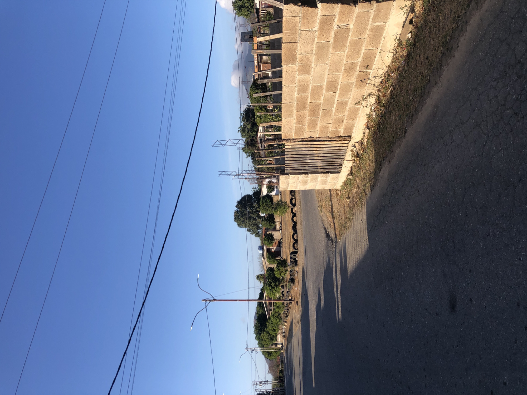 The image depicts a street scene in Altagracia, Venezuela. The road stretches into the distance, bordered by a cinder block wall on the right. A line of trees and utility poles run along the left side of the road. A fence is partially visible behind the cinder block wall. Power lines crisscross the clear blue sky. Shadows are cast on the asphalt road surface. A row of tires lines the roadside, near the trees.