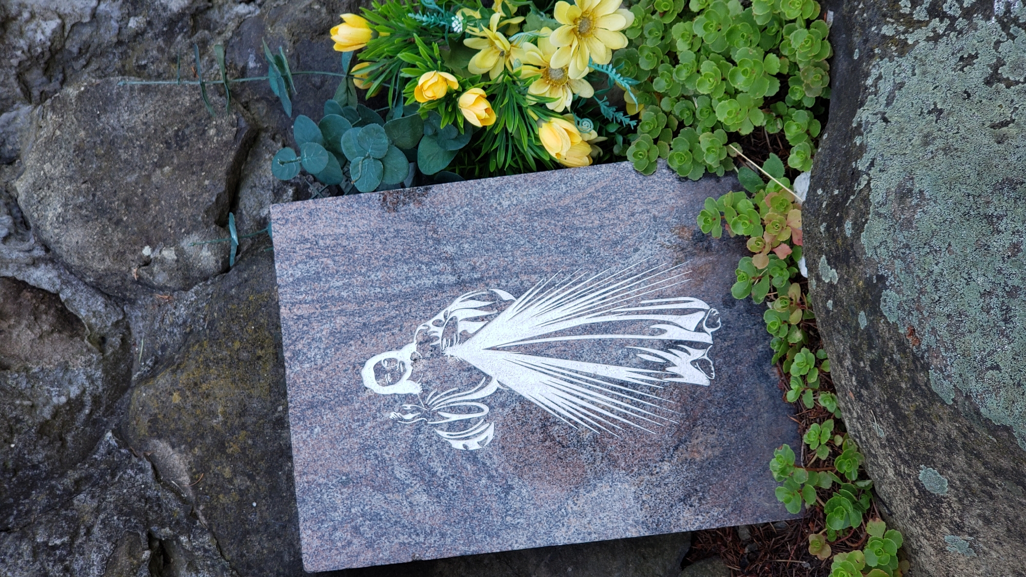 A close-up, eye-level outdoor shot displays a portion of a gravestone adorned with a white silhouette of Jesus Christ, positioned in front of a rough-textured rock wall. To the right of the gravestone, a cluster of artificial yellow flowers and green foliage is arranged. Lush green ground cover plants sprout from the earth and around the base of a large, lichen-covered boulder in the foreground. The scene is likely located in Prešov, Slovakia, given the image's metadata.