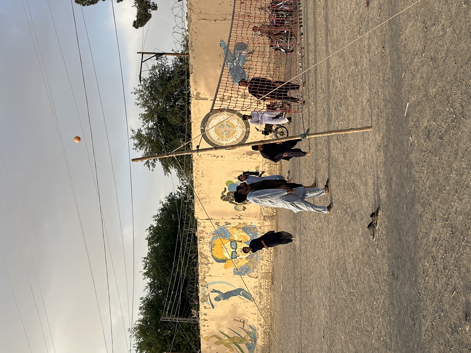 A group of people play volleyball on a dirt court in Quetta City Tehsil, Pakistan. A volleyball net is suspended between two wooden poles. Some players stand, others are in wheelchairs. A yellow volleyball is in the air. A concrete wall forms a backdrop, decorated with graffiti-style paintings. Overhead are electrical wires.