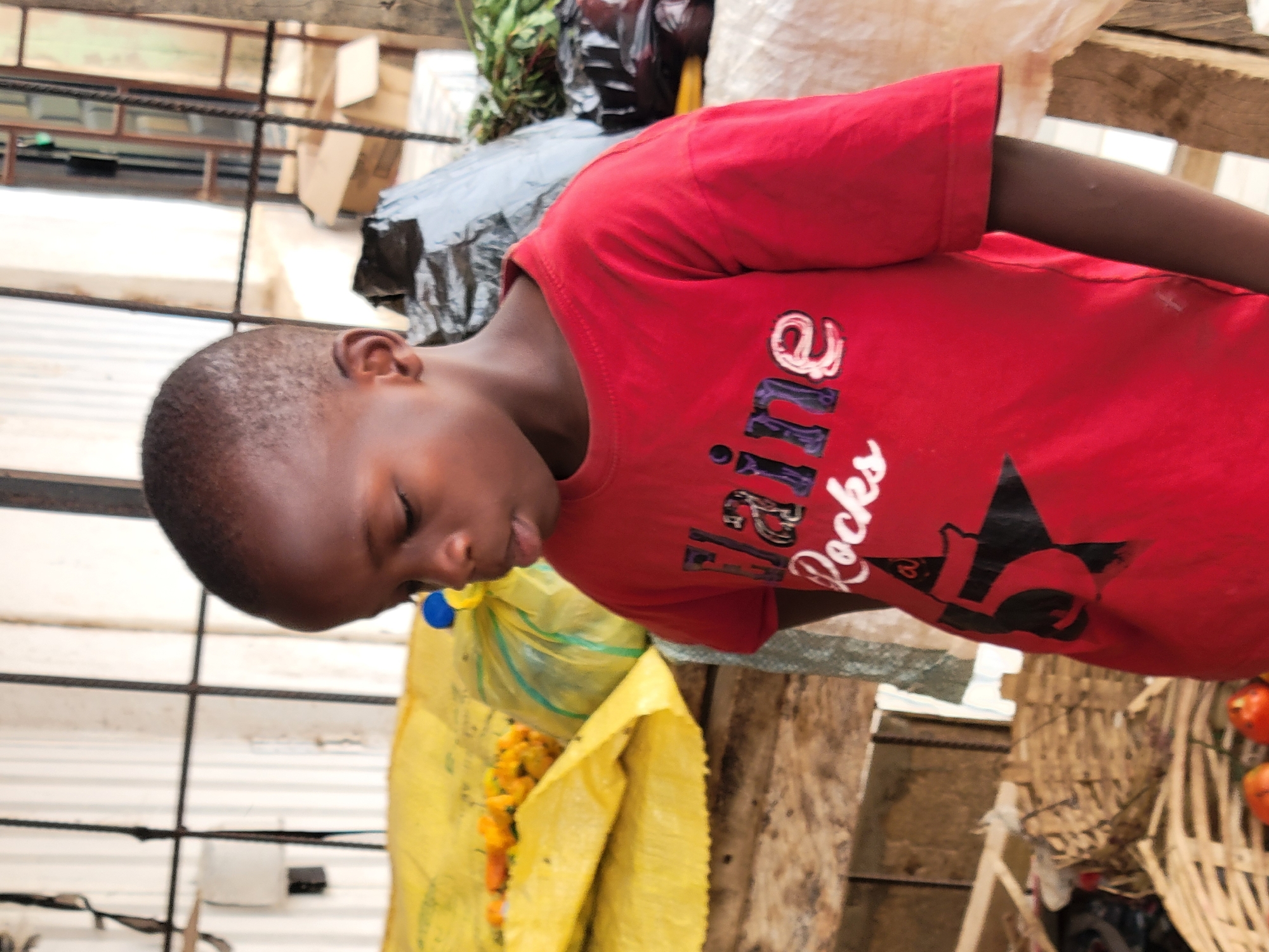 A young child, likely male, is captured in a close-up, side profile shot, resting within what appears to be an outdoor market setting in Jos, Nigeria. The child has dark skin and closely cropped dark hair. His face is turned downwards, with his eyes closed or heavily lidded, suggesting he is either asleep, very tired, or deeply concentrated. His expression appears calm and peaceful.
He is wearing a bright red short-sleeved t-shirt. Adorning the front of the shirt is white cursive text that clearly reads "Elle Rocks," with a large black star graphic partially visible on the lower right side.
The background is a busy and somewhat cluttered outdoor environment, characteristic of a market. Various objects are visible, including large black and white sacks, and a bright yellow plastic bag that seems to be filled with yellow or orange produce. Some green leafy items are visible in another black bag. The structures in the background are composed of informal wooden planks and frameworks, suggesting market stalls, along with corrugated metal sheets forming walls or roofs. On the far right, a woven basket-like container holds several round orange items, possibly tomatoes or small fruits.
The scene is brightly lit, indicative of daytime. The image presents a quiet moment of rest for the child amidst the likely vibrant and bustling atmosphere of a Nigerian market.
