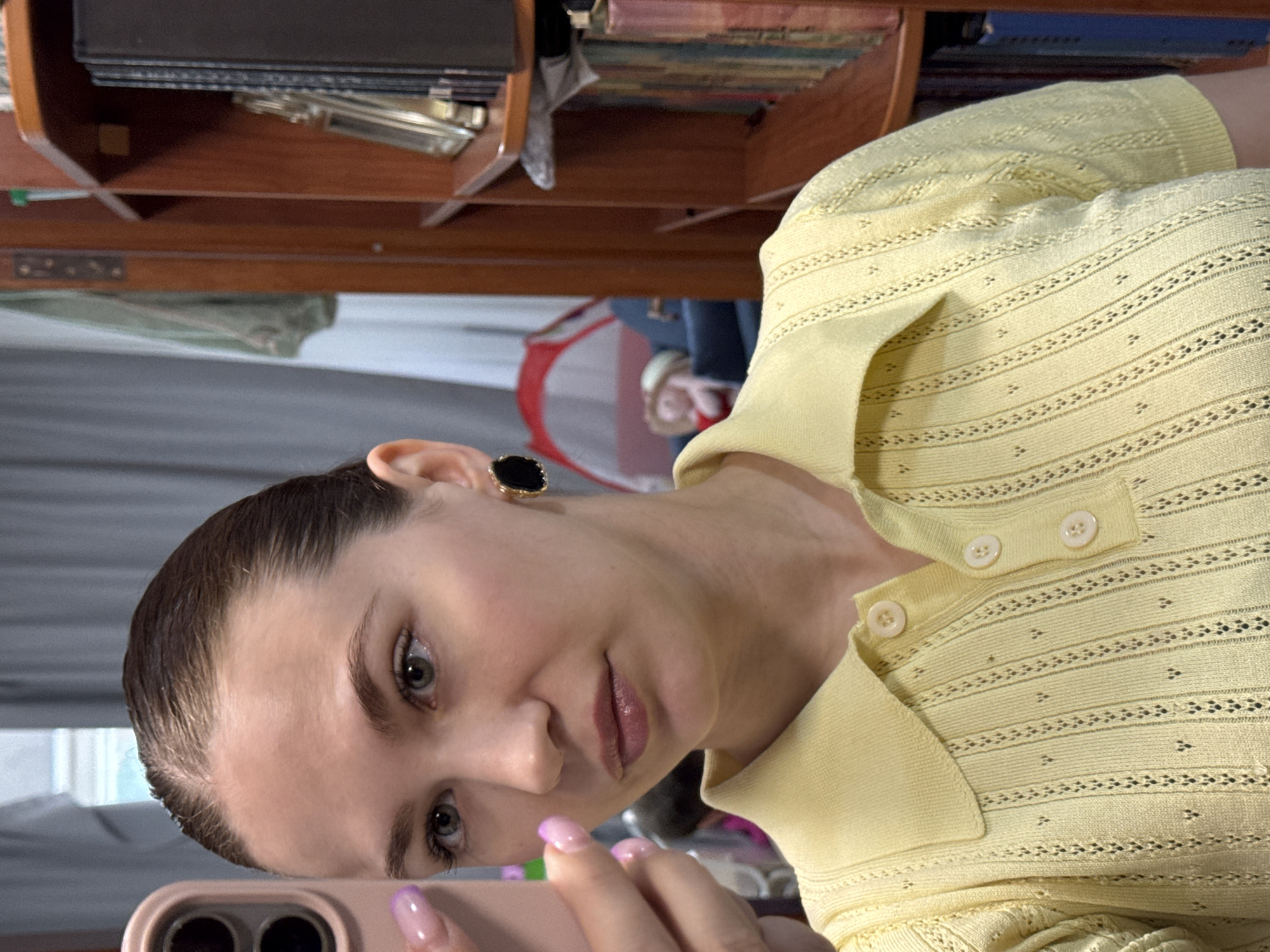 An adult female is depicted in an indoor setting within the city of Chaikovsky, Russia, taking a mirror selfie. She holds a pink smartphone, likely an iPhone, with her left hand, which features an ombre pink-to-purple French manicure. Her light brown hair is slicked back, revealing her forehead. Her eyes are light green/hazel, and she wears minimal makeup, including a sheer lip gloss. A black, possibly onyx, and gold earring is visible on her right ear. She is dressed in a light yellow knitted polo shirt with a patterned, open-work texture and three light-colored buttons on the placket. The background reveals a dark brown wooden bookshelf filled with books and a gold picture frame on the right. To the left, a window with grey and white curtains is visible. Further in the background, a red play tent and a small doll wearing a straw hat seated on a blue object are discernible, indicating a domestic environment.