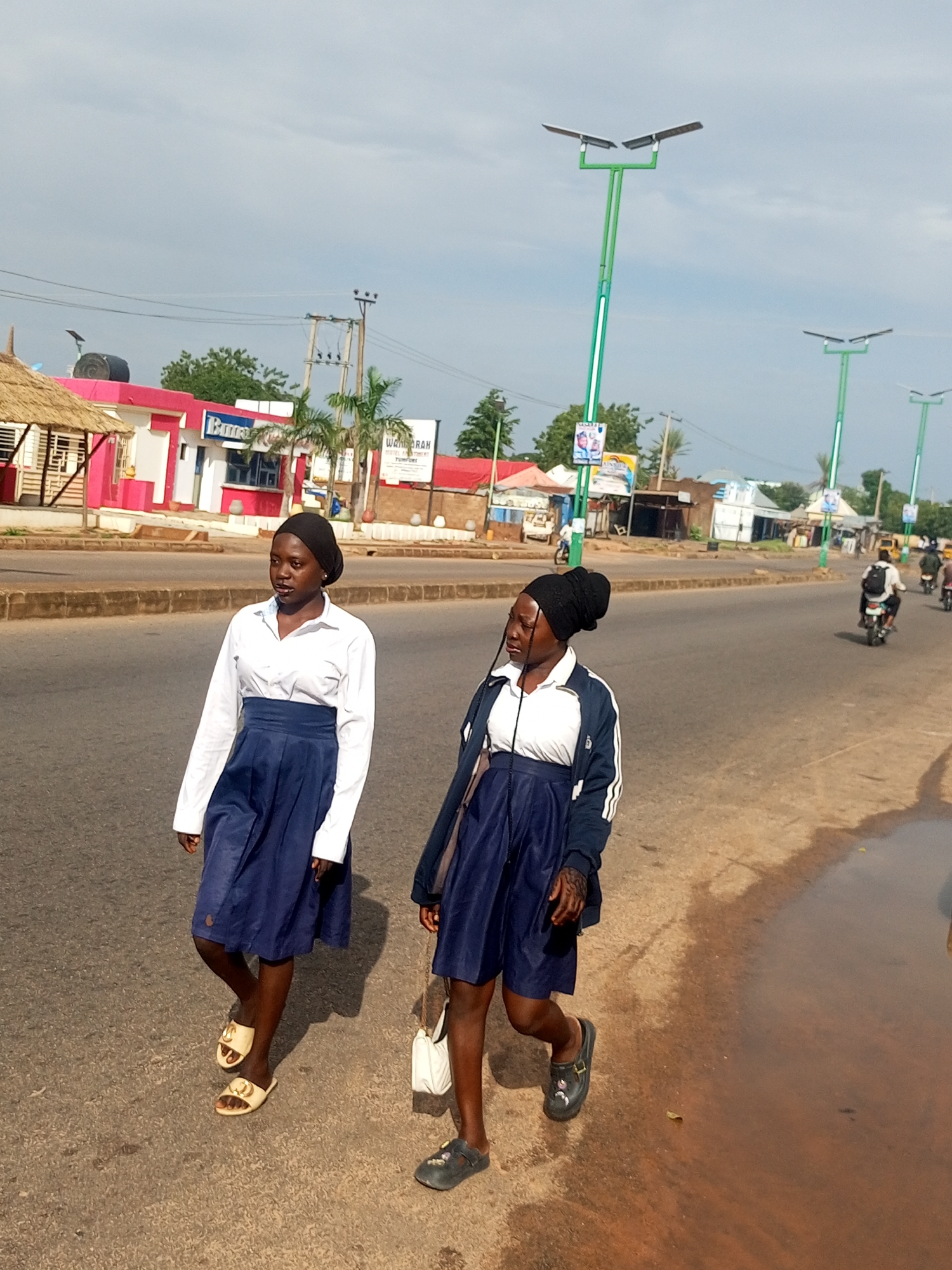 Two young women are walking on a paved road in an outdoor urban environment. The woman on the left is wearing a white long-sleeved shirt, a dark blue pleated skirt, and light-colored slip-on sandals. She has dark hair covered by a black headscarf. The woman on the right is wearing a white shirt, a dark blue jacket with white stripes on the sleeves, and a dark blue pleated skirt. She also has dark hair covered by a black headscarf and is wearing dark slip-on shoes. She is carrying a white handbag over her left shoulder. They appear to be walking in the same direction. The road has a curb and a sidewalk or paved area to the left, with buildings and vegetation visible in the background. There are streetlights with solar panels along the road. Traffic, including motorcycles and a yellow vehicle, is visible in the background. The sky is overcast.