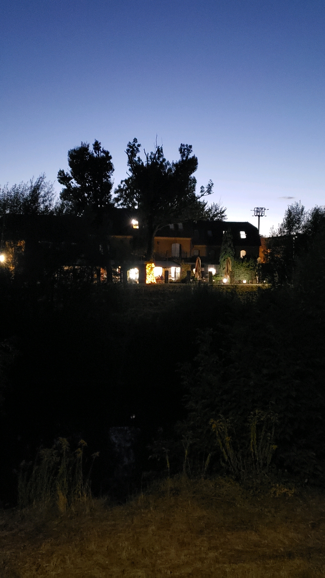 An outdoor scene at dusk features a building with illuminated windows, surrounded by trees. The building appears to be a residential or commercial establishment. The foreground consists of dense vegetation and grass, suggesting a natural or semi-natural environment. The sky is a deep blue, indicative of the twilight hours. There are no people visible in the frame. The location is identified as Prešov, Slovakia.