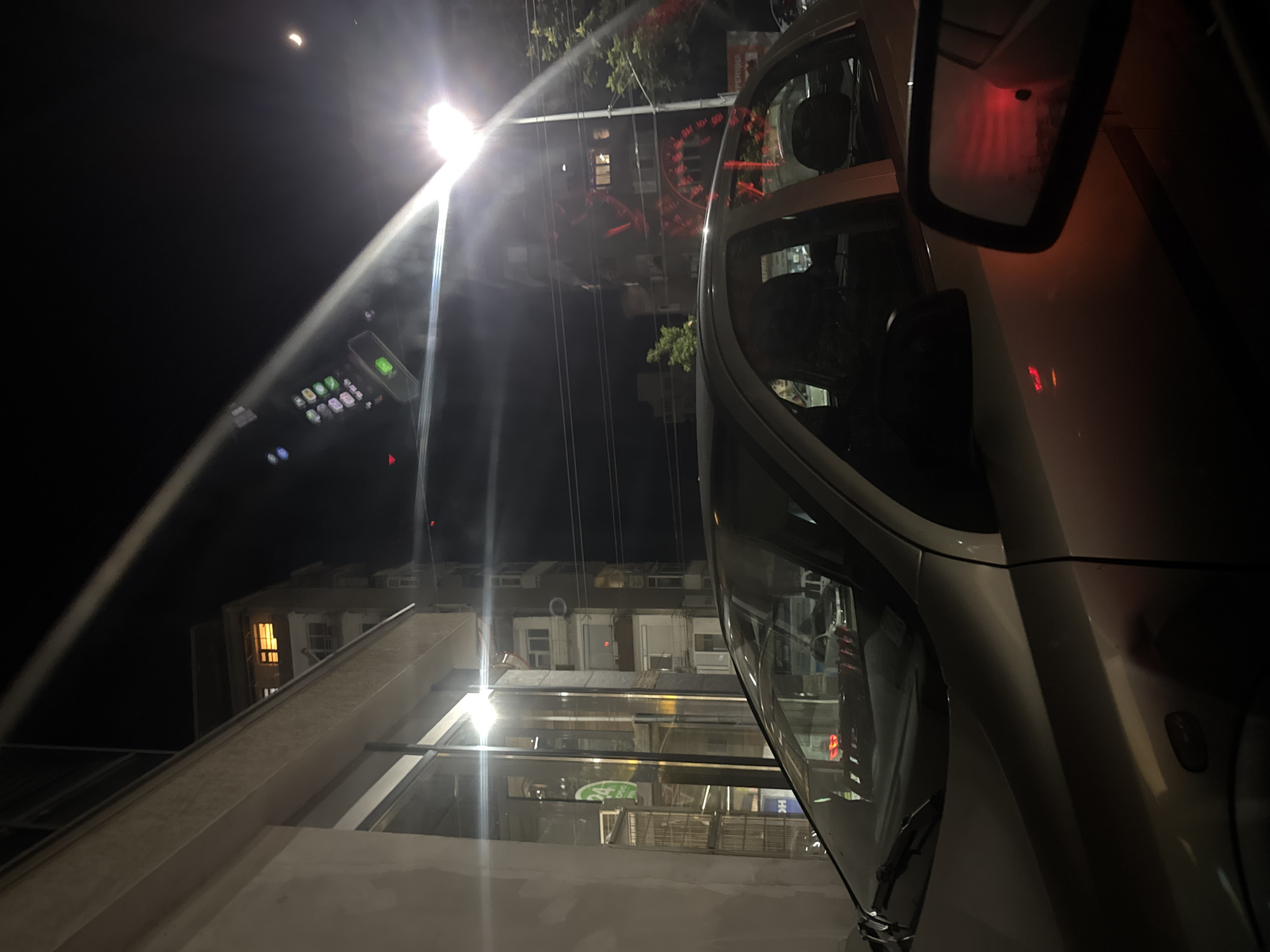 Captured at night, the image presents a view from within a vehicle, likely in Stara Zagora, Bulgaria. The foreground is dominated by a car, with parts of its interior and exterior visible. A side mirror reflects a faint red glow. Through the car windows, the cityscape emerges, featuring a building facade with lighted windows, suggesting residential or commercial occupancy. Streetlights cast a bright glare, obscuring some details and creating lens flare effects. Wires stretch across the skyline, typical of urban infrastructure. The setting appears to be a street scene, potentially near a parking area or roadway. A crescent moon is visible in the upper left corner of the image.