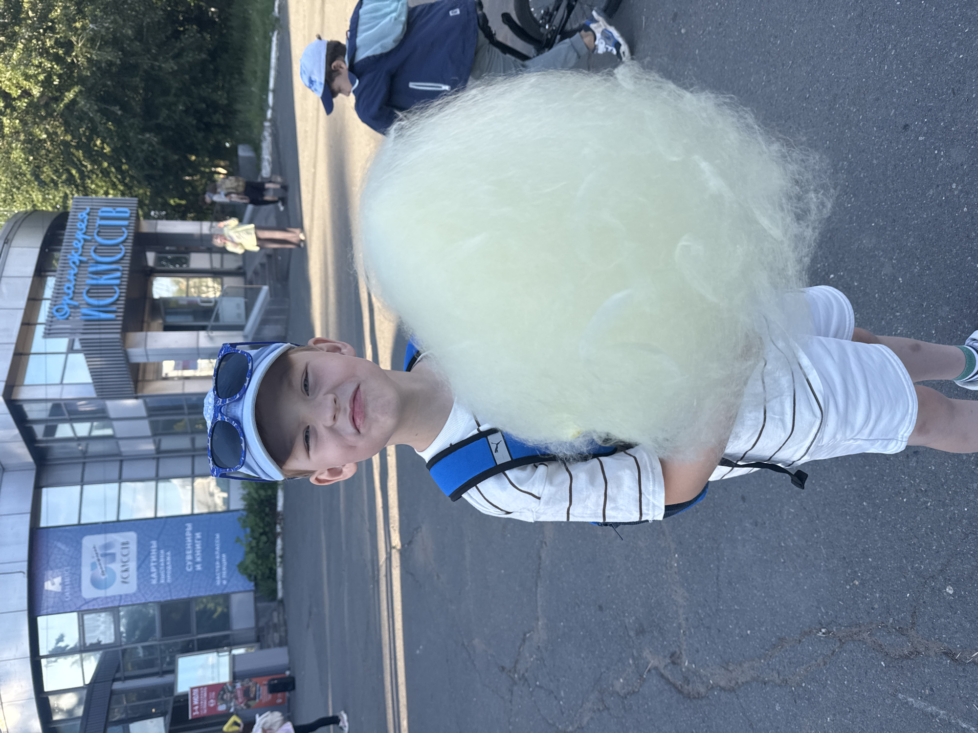 A young boy with a light complexion, wearing a light blue baseball cap with dark sunglasses resting on its brim, a white striped t-shirt with a visible Puma logo, white shorts, and a blue backpack, stands centrally in the frame, holding a large mass of light yellow/white cotton candy in both hands. He presents a smiling or puckered facial expression, with visible dimples. His lower legs show green and white striped socks and white sneakers.
The scene unfolds outdoors on dark asphalt pavement, showing cracks and minor debris, under bright sunlight casting distinct shadows. In the background, a modern building with a glass and metallic facade is visible. Prominently displayed on the building is blue Cyrillic signage reading "Оранжерея ИСКУССТВ," translating to "Greenhouse of Arts," identifying the location as a cultural center in Izhevsk, Russia. A large banner on the left portion of the building also displays Cyrillic text including "ИСКУССТВ" (Arts), "КАРТИНЫ" (Paintings), "ВЫСТАВКИ" (Exhibitions), "ПРОДАЖА" (Sale), "СУВЕНИРЫ" (Souvenirs), "И КНИГИ" (And Books), "МАСТЕР-КЛАССЫ" (Master Classes), and "И ЛЕКЦИИ" (And Lectures). A smaller sign near the entrance indicates "3-4 ИЮЛЯ" (3-4 July).
To the right of the central boy, another boy in a white baseball cap, dark blue jacket, and grey pants is visible from the back, on a bicycle with blue and white sneakers. Near the building's entrance, two adults are present; one in a light-colored long-sleeved shirt and light brown pants, appearing to gesture, and another partially obscured behind them. The far left of the frame shows the lower legs and feet of a person walking away. Green foliage and trees are visible on the right side of the background, behind the building structure.