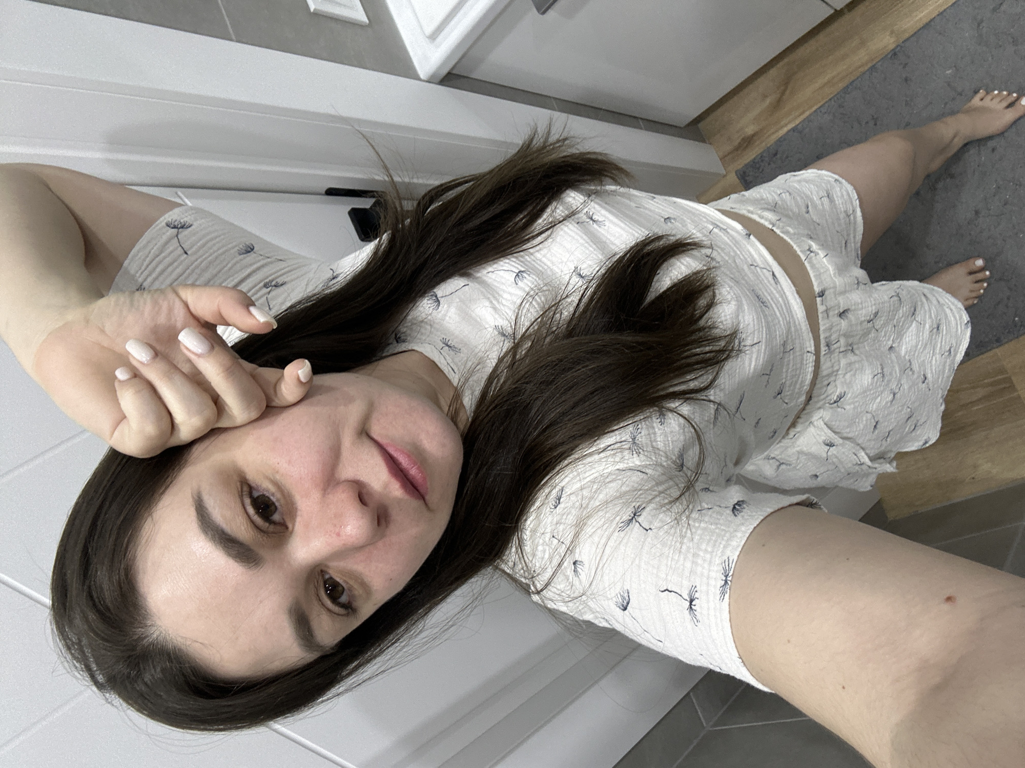 A selfie-style image shows a woman indoors, possibly in a bathroom based on the tiles and fixtures visible in the background. She is wearing a light-colored, patterned pajama set. Her dark hair is loose. The woman's right hand is near her face. Her toes are painted white, and she is standing on a gray bathmat. The location in the city of Chaikovsky, Russia, cannot be determined from the image.