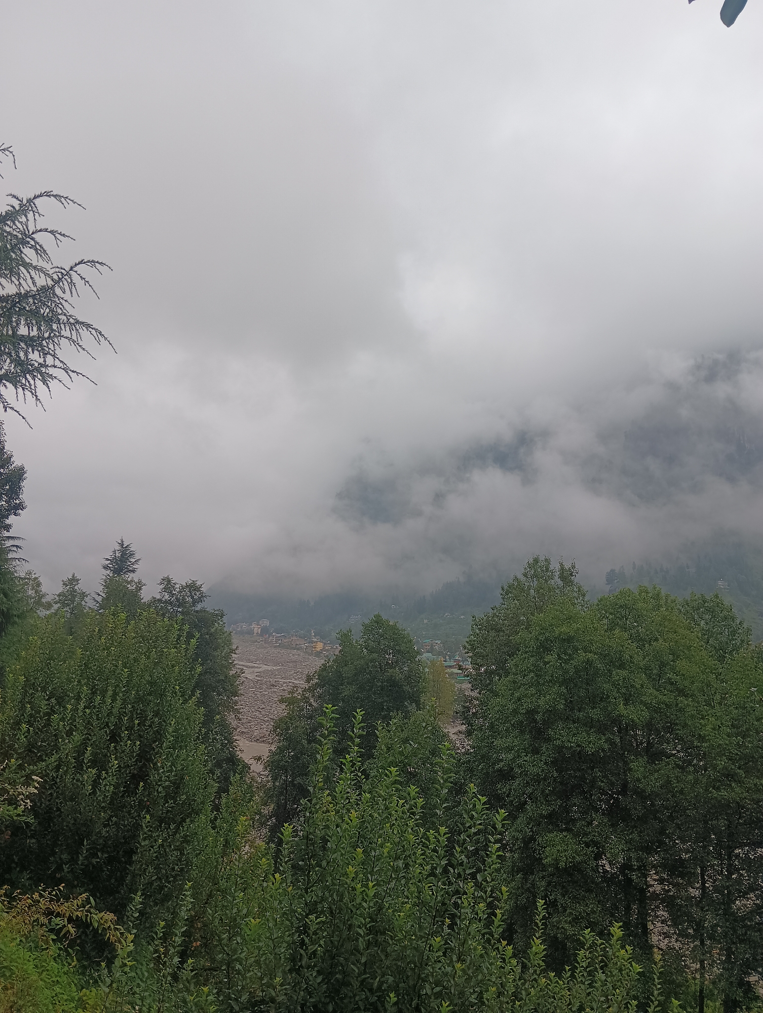 A vertical, eye-level, full shot captures a misty mountain landscape with a village nestled in the valley. The foreground is dominated by lush green trees and foliage. In the mid-ground, a wide, rocky riverbed is visible, with a small village of multi-story buildings situated along its banks. The background is shrouded in thick, grey clouds that obscure the mountain peaks, giving the scene a somber, overcast atmosphere. The overall impression is one of a remote, tranquil, and somewhat melancholic mountain village enveloped in mist. The location is identified as Goshal, India.