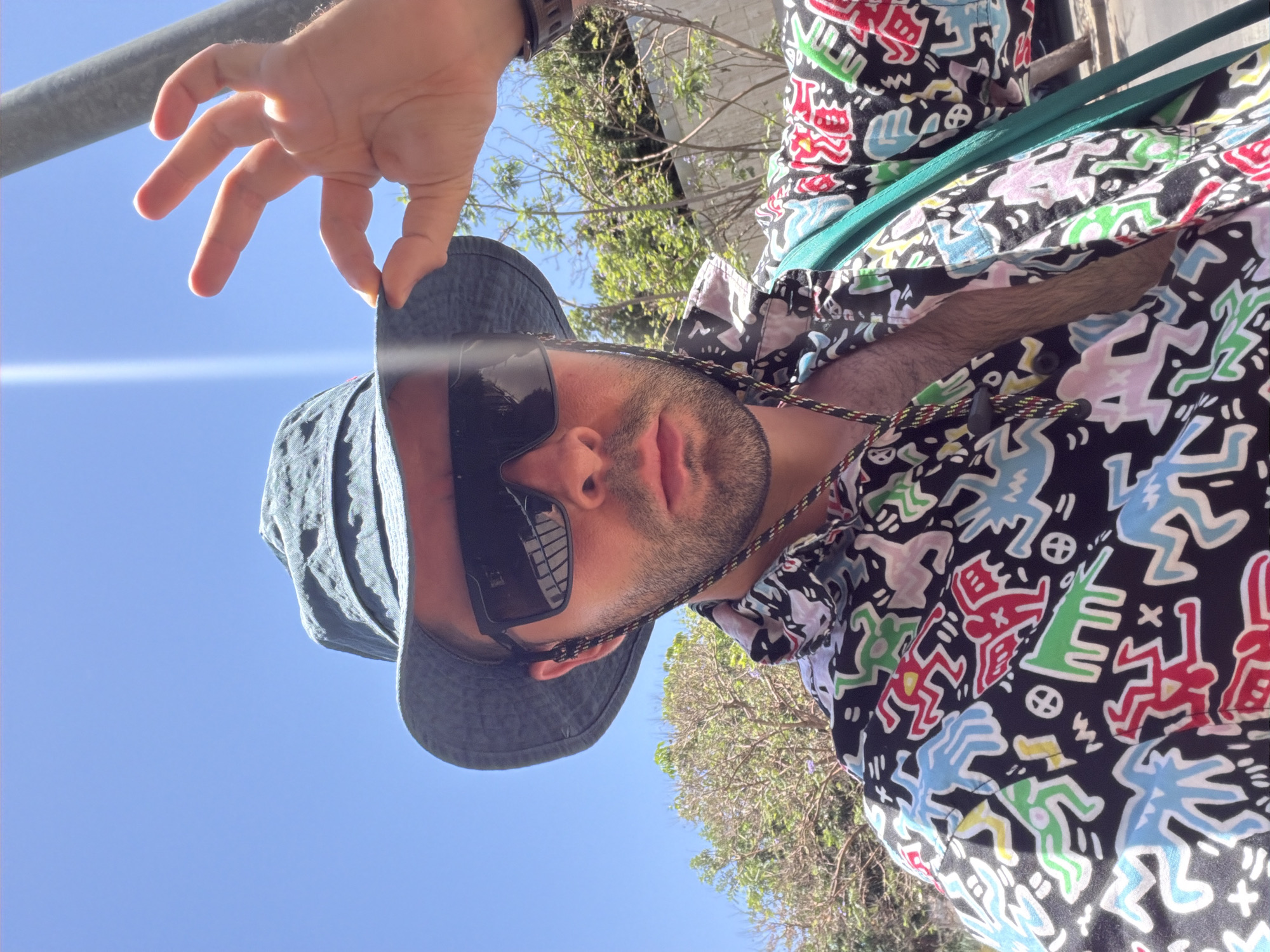 A male individual is captured in a sunlit outdoor selfie, likely in Tel-Aviv, Israel. He wears a dark blue bucket hat, black rectangular sunglasses that reflect a bright vertical sun flare and a grid-like pattern, and a black short-sleeved collared shirt adorned with a vibrant, multi-colored pattern featuring abstract human-like figures in the style of Keith Haring. The shirt is unbuttoned, revealing chest hair, and a colorful, braided cord with a black stopper bead hangs around his neck. A teal or green strap, presumably from a bag, crosses his upper torso on the right side. A dark wristwatch is visible on his right wrist. His right hand is raised, holding the brim of his bucket hat. The background features a clear, bright blue sky, partially bare trees with sparse green foliage and hints of purple blossoms, and a tall grey pole on the right. The strong sunlight casts bright reflections and creates a lens flare.