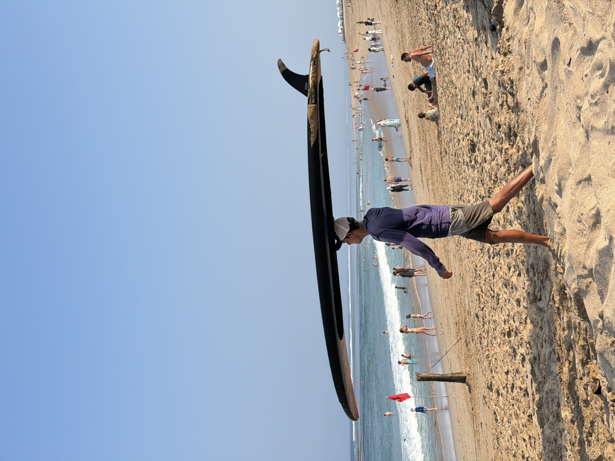 An eye-level, medium shot captures a man walking along a sandy beach, carrying a black surfboard horizontally over his head. He is wearing a light-colored cap, a long-sleeved purple shirt, and gray shorts. The man is positioned in the foreground, slightly off-center to the right. Behind him, the beach is populated with numerous beachgoers. Some are wading in the ocean, while others are sitting or standing on the sand. The ocean in the background displays gentle waves and is a clear blue. The sky above is a bright, cloudless blue. A red flag is visible on a wooden pole near the water's edge. The sand is light brown and appears dry and rippled in the foreground.