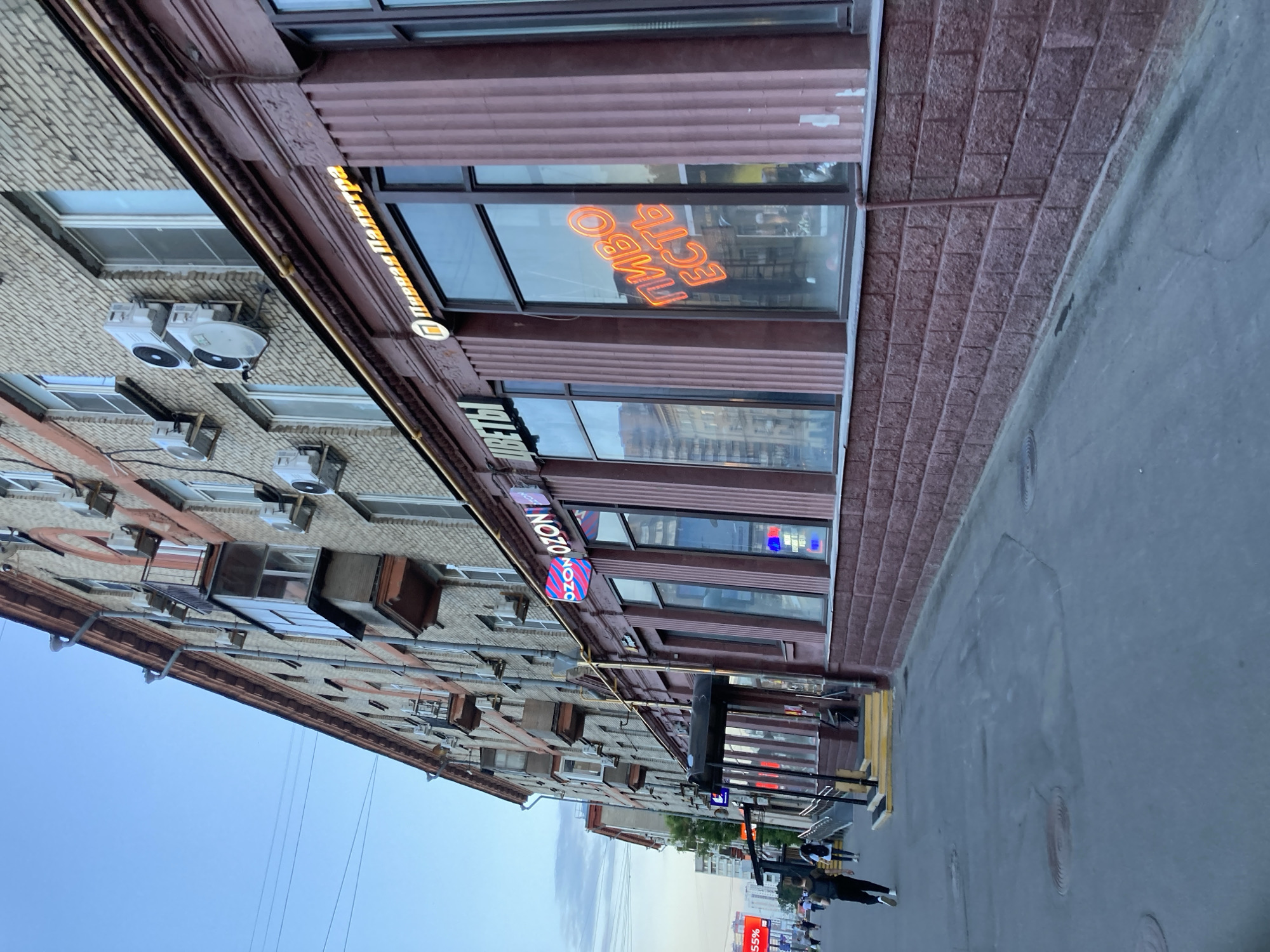 This is a medium-angle, eye-level shot of a building facade in Khimki, Russia, with a sidewalk in the foreground. The building is a multi-story apartment building with brick and stone elements. The ground floor features several storefronts with large windows. One storefront has a neon sign in Russian that reads "ПИВО ЕСТЬ" (Pivo Est), meaning "Beer is Here." Another storefront has a neon sign for "OZON." There are also signs for "ЦВЕТЫ" (Tsvety), meaning "Flowers." Several air conditioning units are visible on the building's facade above the storefronts.
On the sidewalk, a few people are walking. One person in the foreground is walking away from the camera, wearing a black backpack. The sidewalk is made of asphalt and shows some wear and tear, with several manhole covers visible. The sky above is a clear blue with a few wispy clouds. The overall lighting suggests it is late afternoon or early evening. The scene depicts a typical urban streetscape in a Russian city.