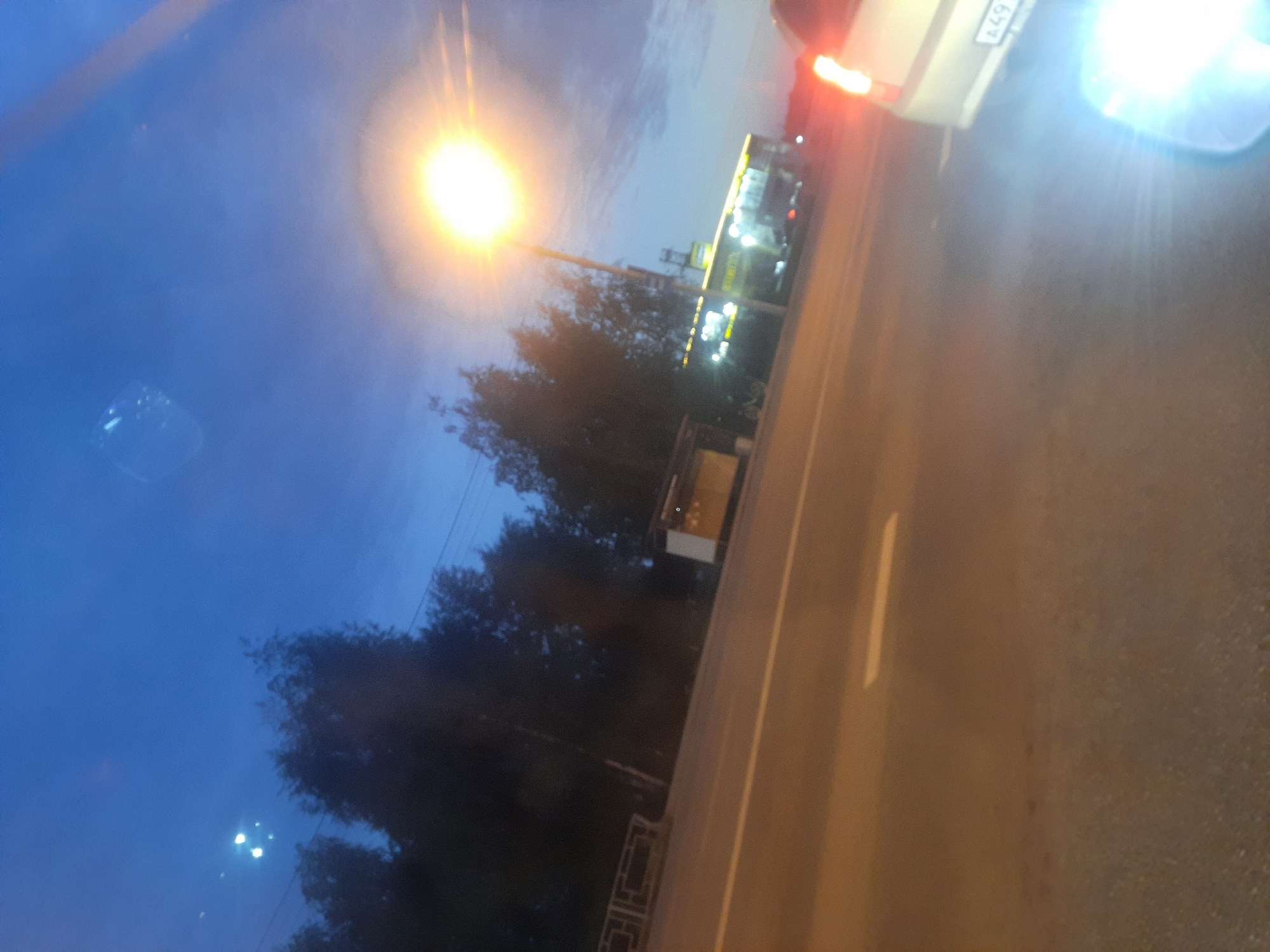 The image captures a nighttime street scene in Barnaul, Russia, likely during dusk. The sky is a deep blue, transitioning into twilight, with a bright, possibly artificial light source illuminating the upper right quadrant of the frame. Silhouetted trees line the left side of the road, and a guardrail is visible in the lower left. A road with a white lane marker stretches into the distance. On the right side of the road, there's a building with illuminated windows, possibly a shop or a bus stop, and a street lamp with a very bright light. A vehicle, with its taillights illuminated and license plate visible, is positioned in the lower right corner, facing away from the camera. The overall impression is that of a dimly lit urban environment at the end of the day.