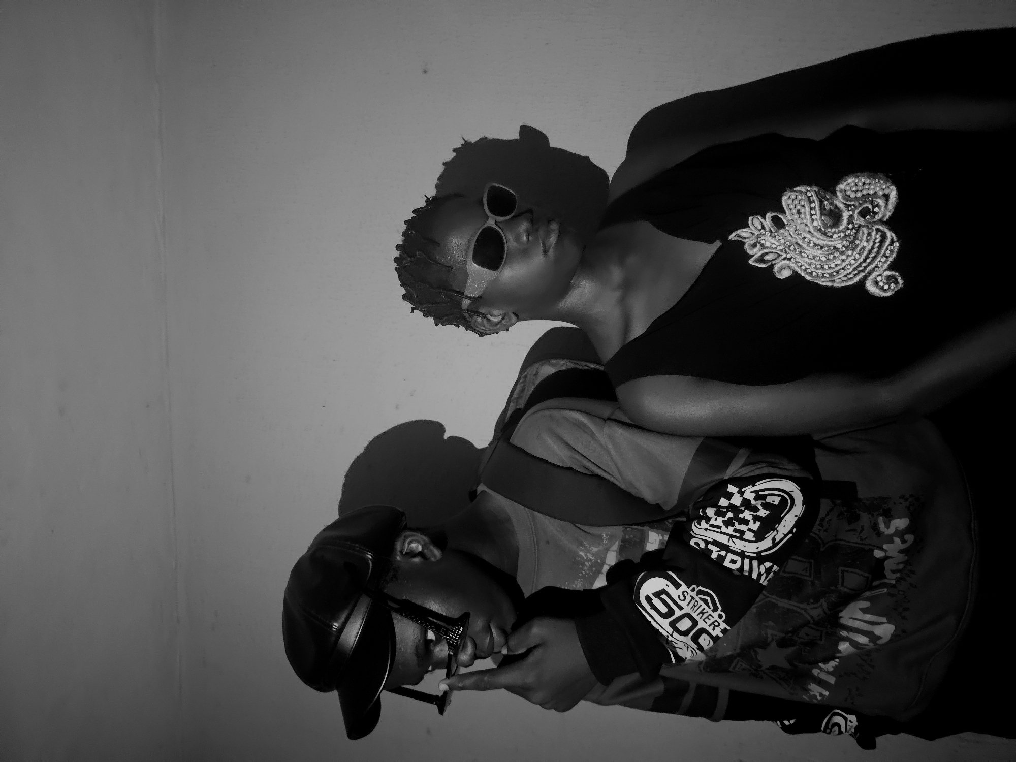 Two individuals are captured in a monochrome image against a plain wall. The person at the bottom wears a cap, glasses, and a patterned jacket, appearing to adjust their glasses. The upper individual is seen with sunglasses and an embellished top. The location is presumed to be Nkwelle, Nigeria, based on the prompt. There are no clear indicators of the environment beyond the wall and minimal shadows cast by the subjects. The interaction is ambiguous, with no apparent physical contact or communication.
