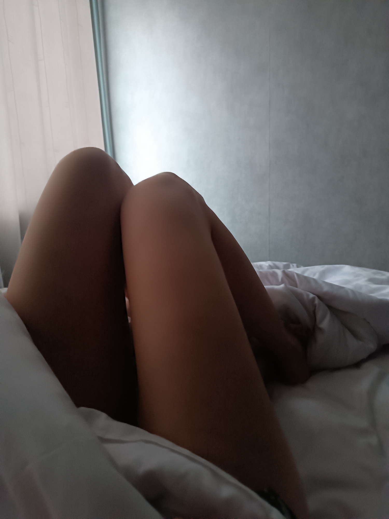 The image shows a pair of legs resting on a white bed. A white curtain is visible in the background, along with a gray wall. The location is a room with a bed.