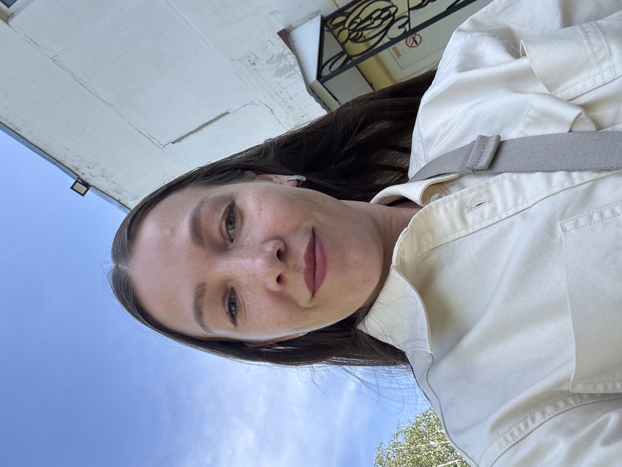 A woman with light skin and dark hair is looking at the camera. She has green eyes, freckles, and a slight smile. She is wearing a light-colored shirt with a gray strap over her shoulder. The sky is blue in the background. The location appears to be in Chaikovsky, Russia, as there is a building with a sign that appears to have Cyrillic writing on it.