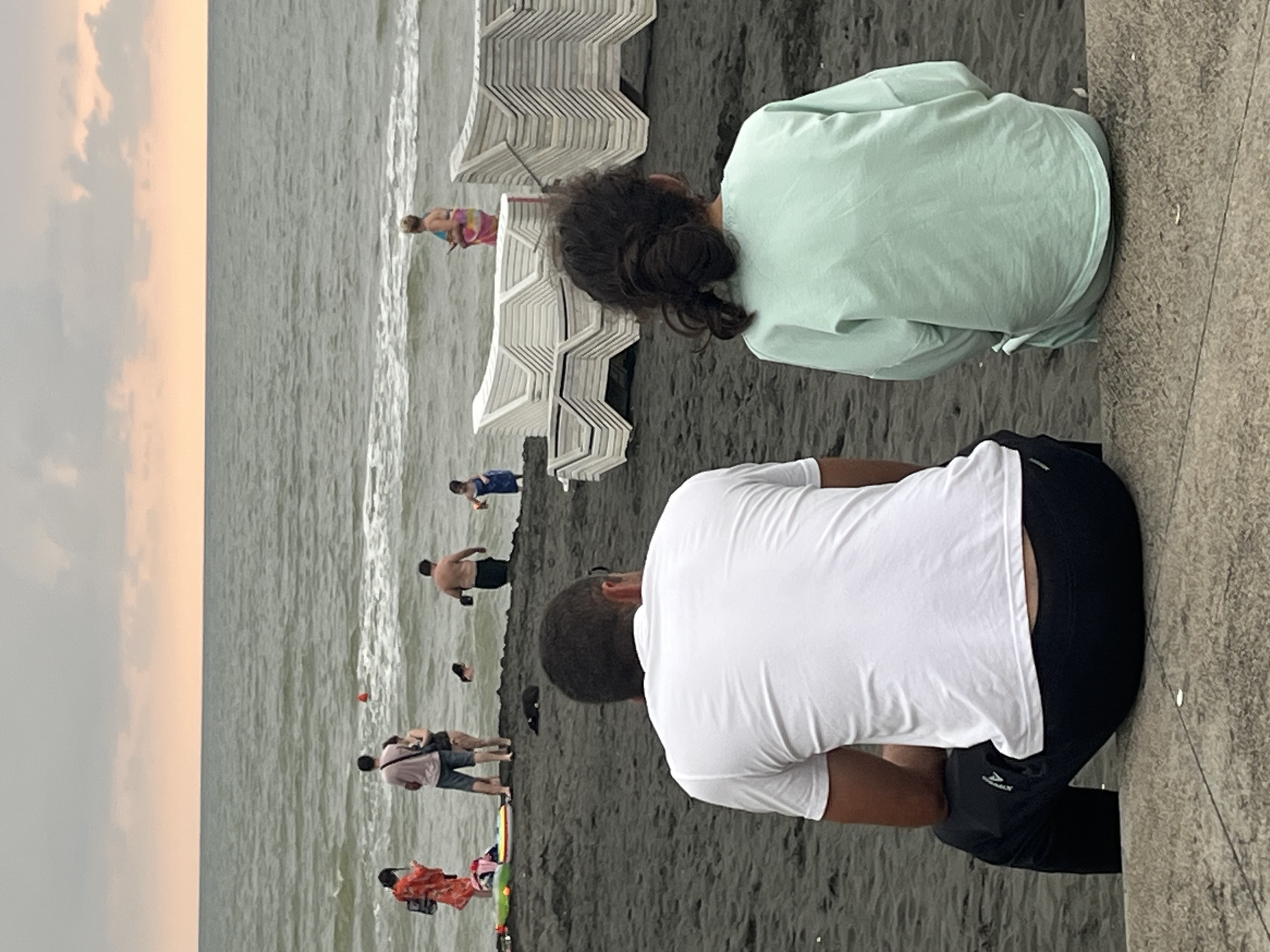 The image depicts a beach scene in Ureki, Georgia, with the Black Sea in the background under an overcast sky. In the foreground, a man wearing a white t-shirt and black pants is seated with his back to the camera, as is a young girl wearing a mint green long-sleeved shirt. The sand is dark, and several stacks of white lounge chairs are visible further back on the beach. In the middle ground, a group of people are near the water. A woman in a bright orange garment stands on the left, and a man and woman in swim attire are near the water's edge. Other individuals are in the sea, including a man with his torso visible and a child. The water is choppy, and waves are breaking on the shore. The overall atmosphere appears relaxed and typical of a beach day.