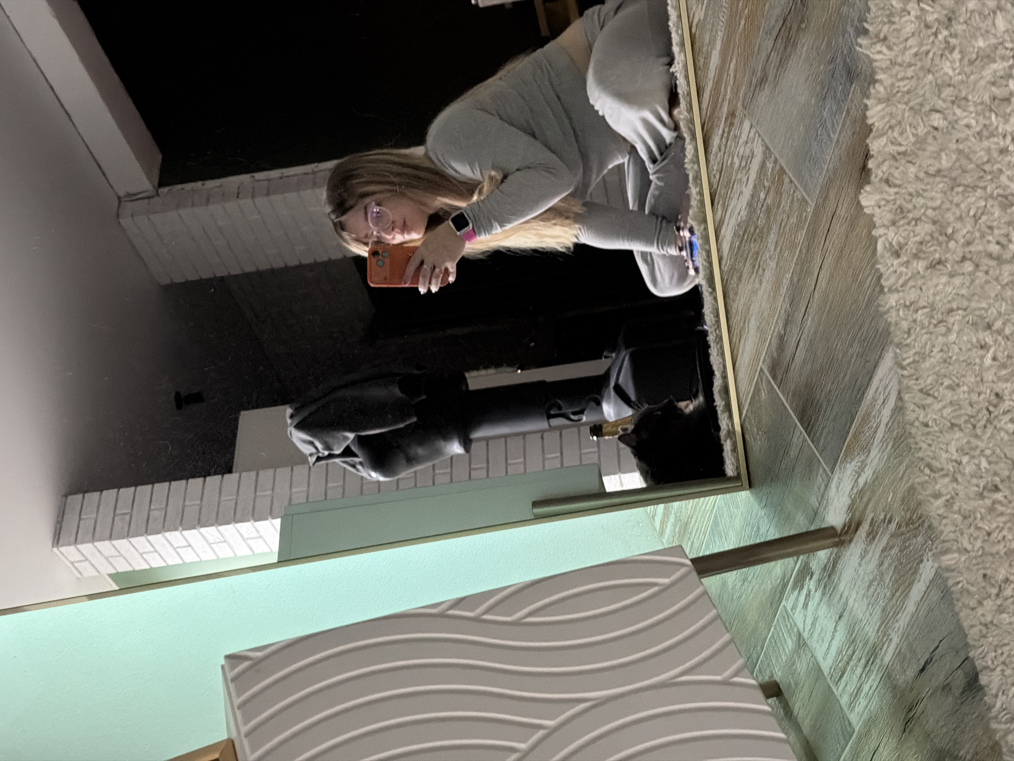 A young woman, reflected in a mirror, is holding a cell phone, presumably taking a selfie. She has long blond hair and wears glasses, a gray sweatsuit, and a pink smartwatch. The mirror reflects a portion of a living space with white-painted brickwork, a dark sofa, and a side table with a patterned front. The flooring is a light-colored wood-look tile, bordered by a shaggy rug. The location is unknown and cannot be determined from the image.